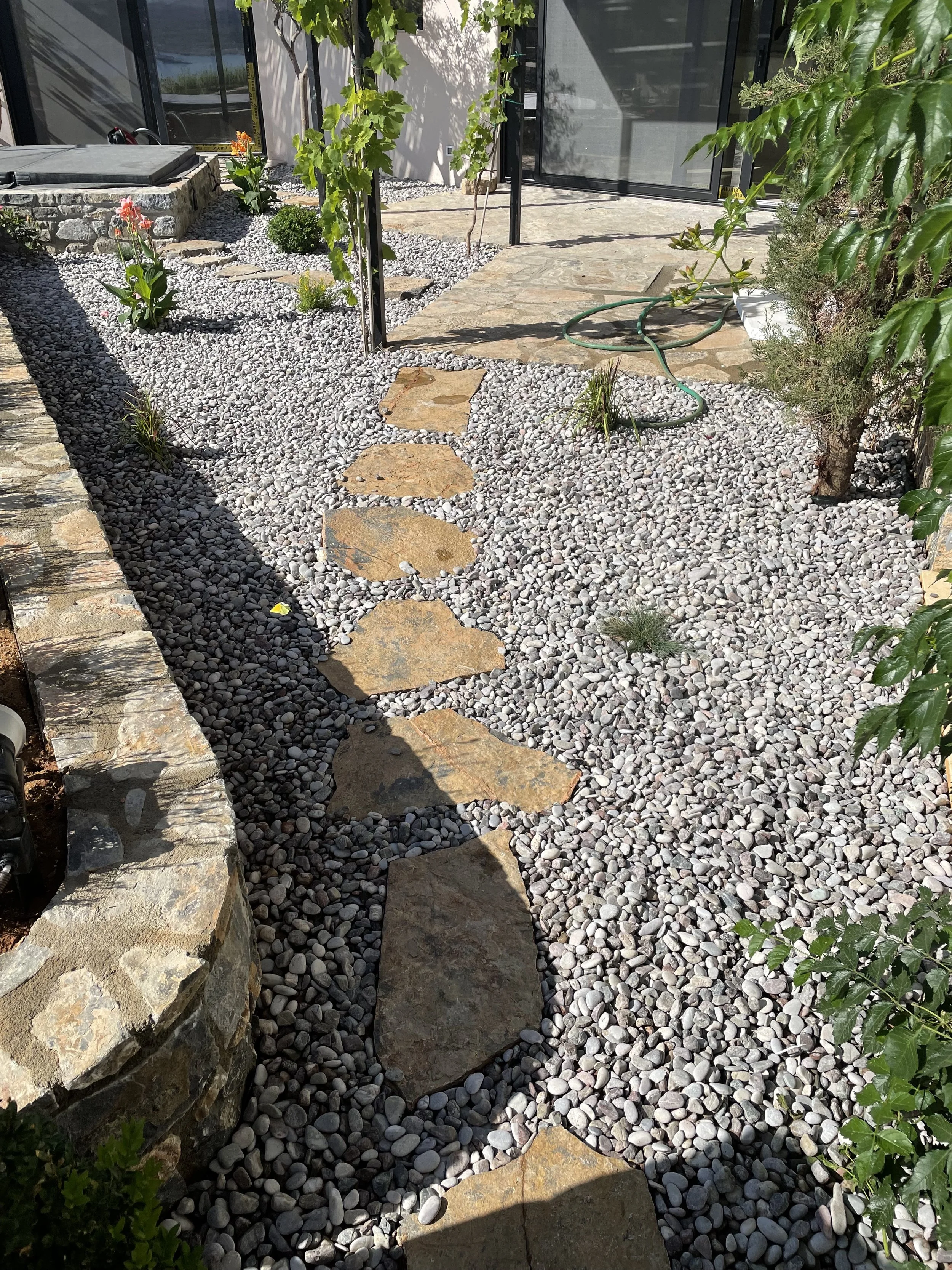 A small garden pathway made of irregular stone slabs surrounded by light gray pebbles, with some green plants and bushes along the sides, and a black metal frame structure in the background.