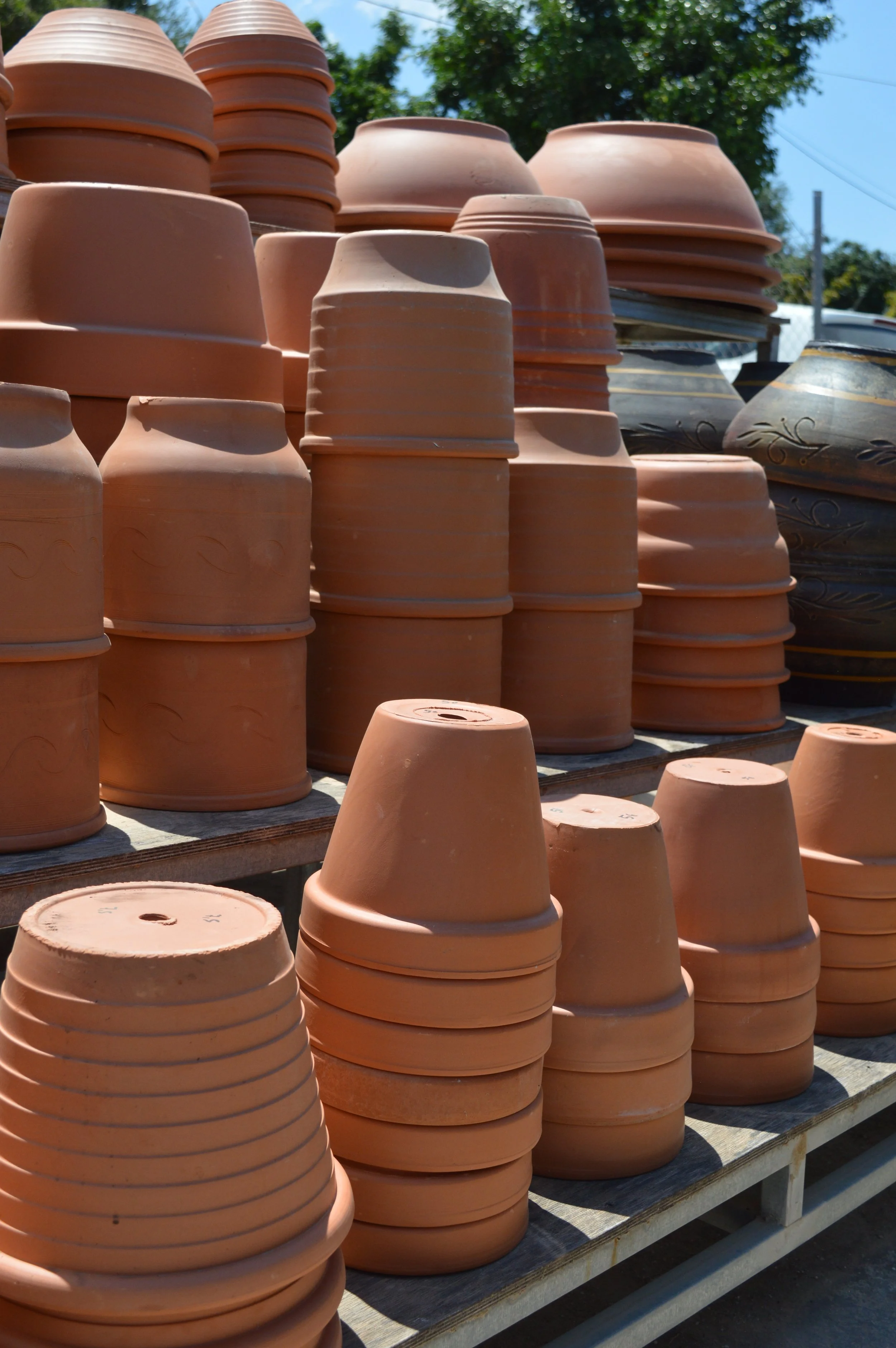 Stacks of unglazed terra cotta flowerpots arranged on wooden shelves outdoors, with a background of green trees and a blue sky.