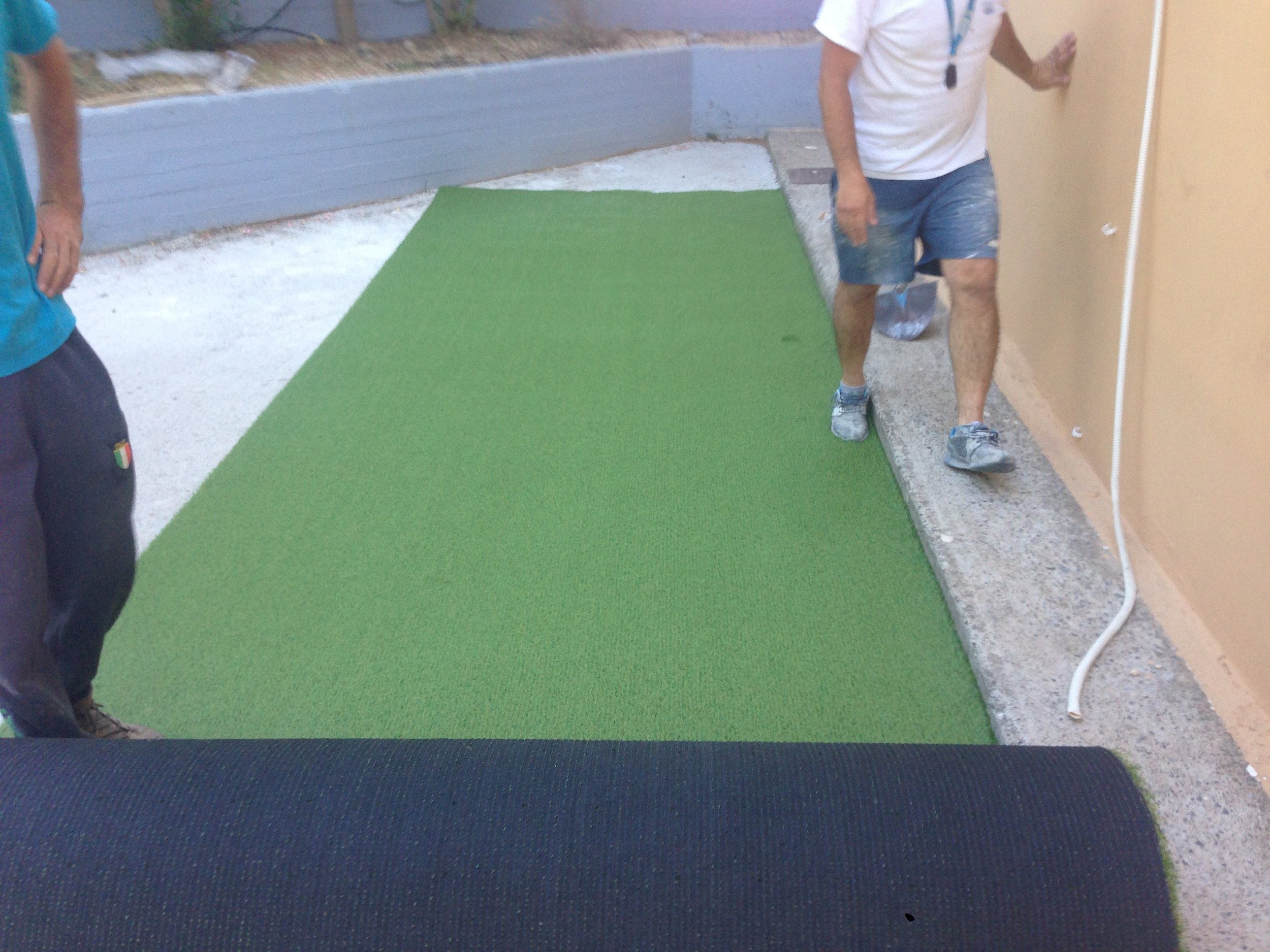 Outdoor scene showing two people near a green artificial turf, with one person partially visible on the left and another person walking on the turf wearing shorts and a white shirt. The area appears to be part of a construction or renovation site wit