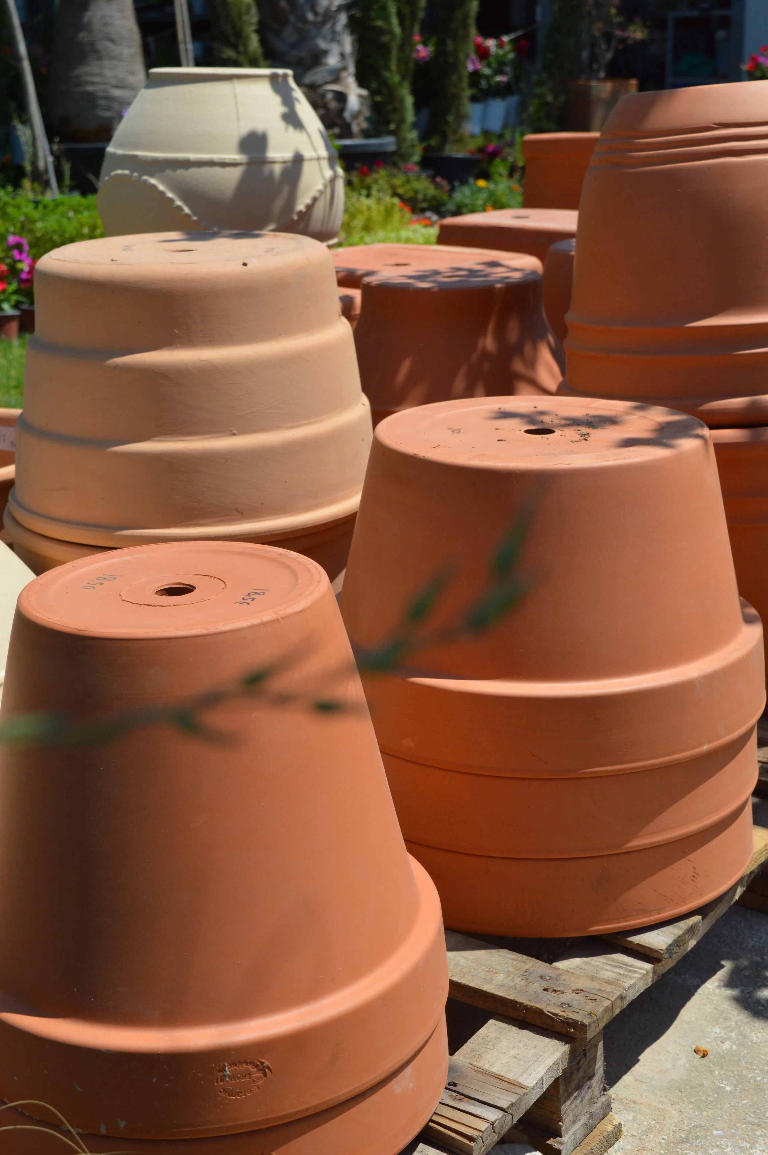 Multiple large terracotta plant pots stacked and placed on wooden pallets outdoors, with a garden and flowers in the background.