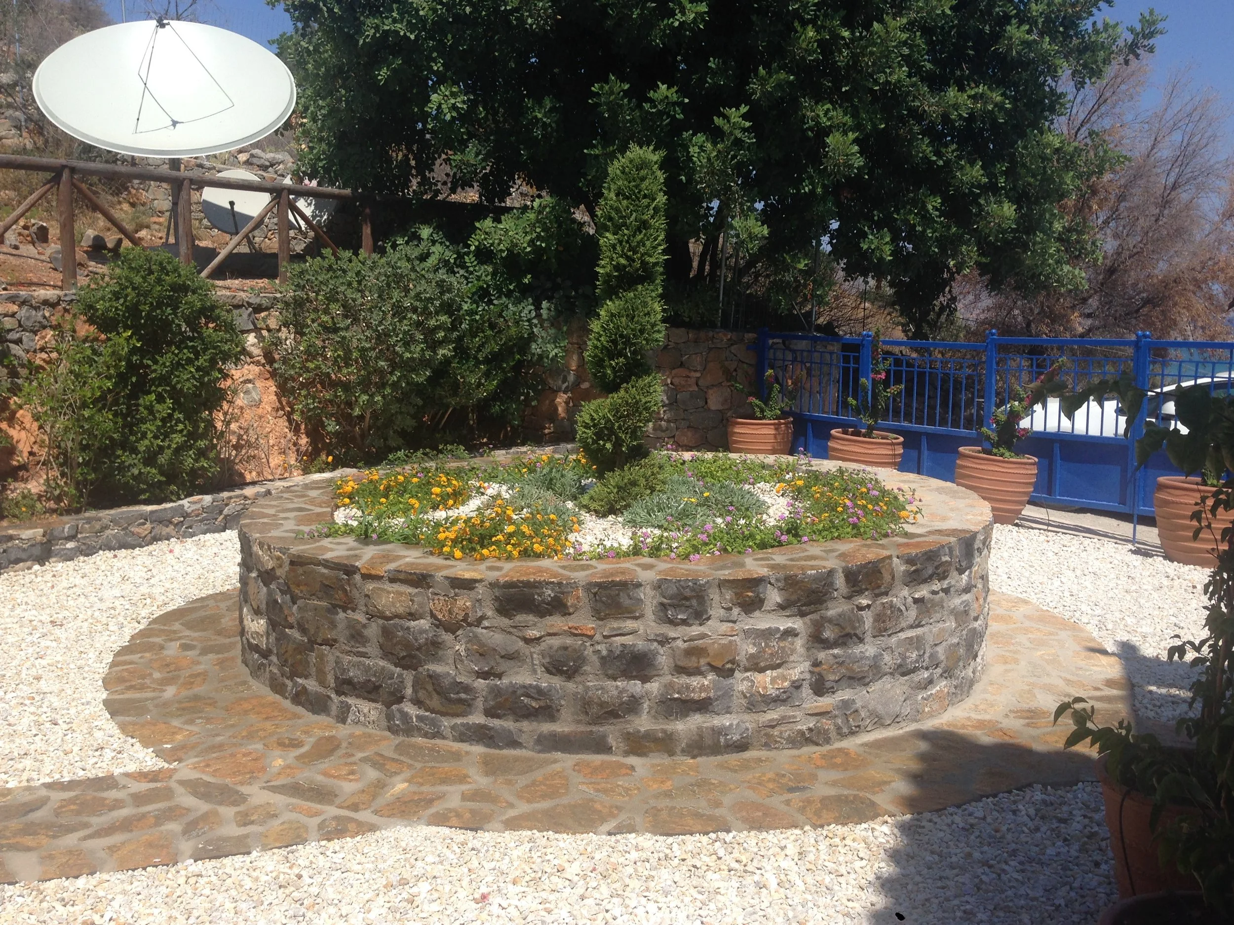 A garden scene with a stone circular flower bed filled with yellow, purple, and white flowers, surrounded by gravel. There is a large tree with green leaves, several potted plants, a blue painted fence, and a satellite dish mounted on a pole in the b