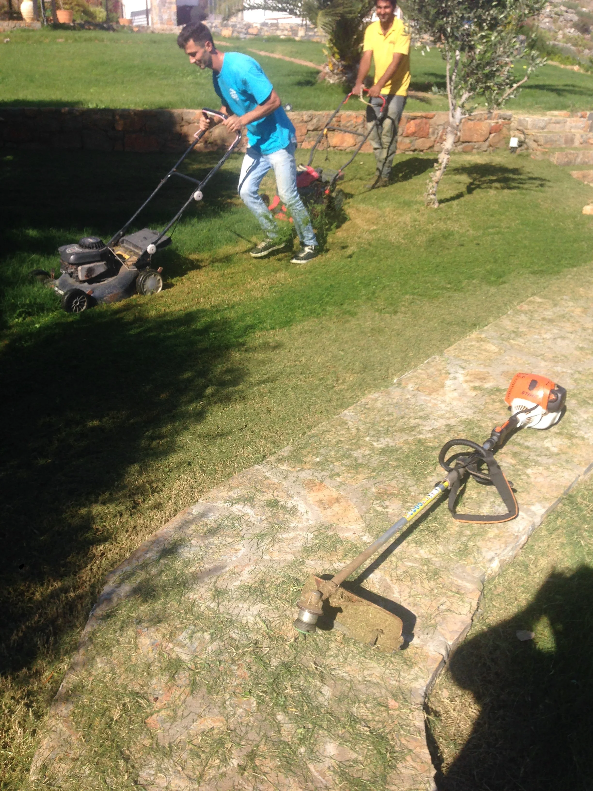 Two men are mowing the lawn outdoors. One man is pushing the lawnmower, and another man is standing behind him. There are trees, a stone border, and a grassy area in the background.