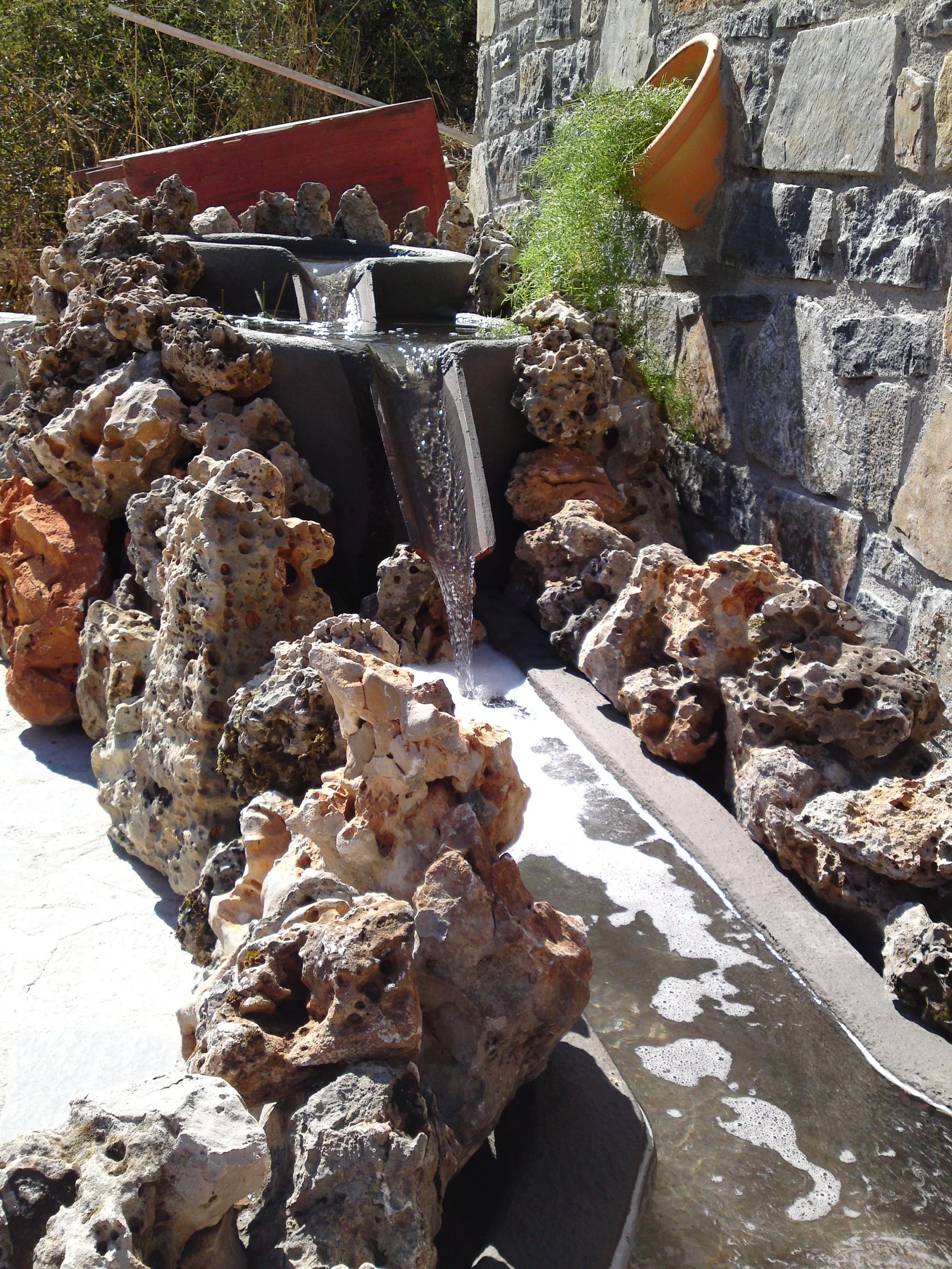 A small water fountain with water flowing over black stone into a basin, surrounded by rocks and a stone wall. A yellow flower pot with green plant is on the wall to the right.