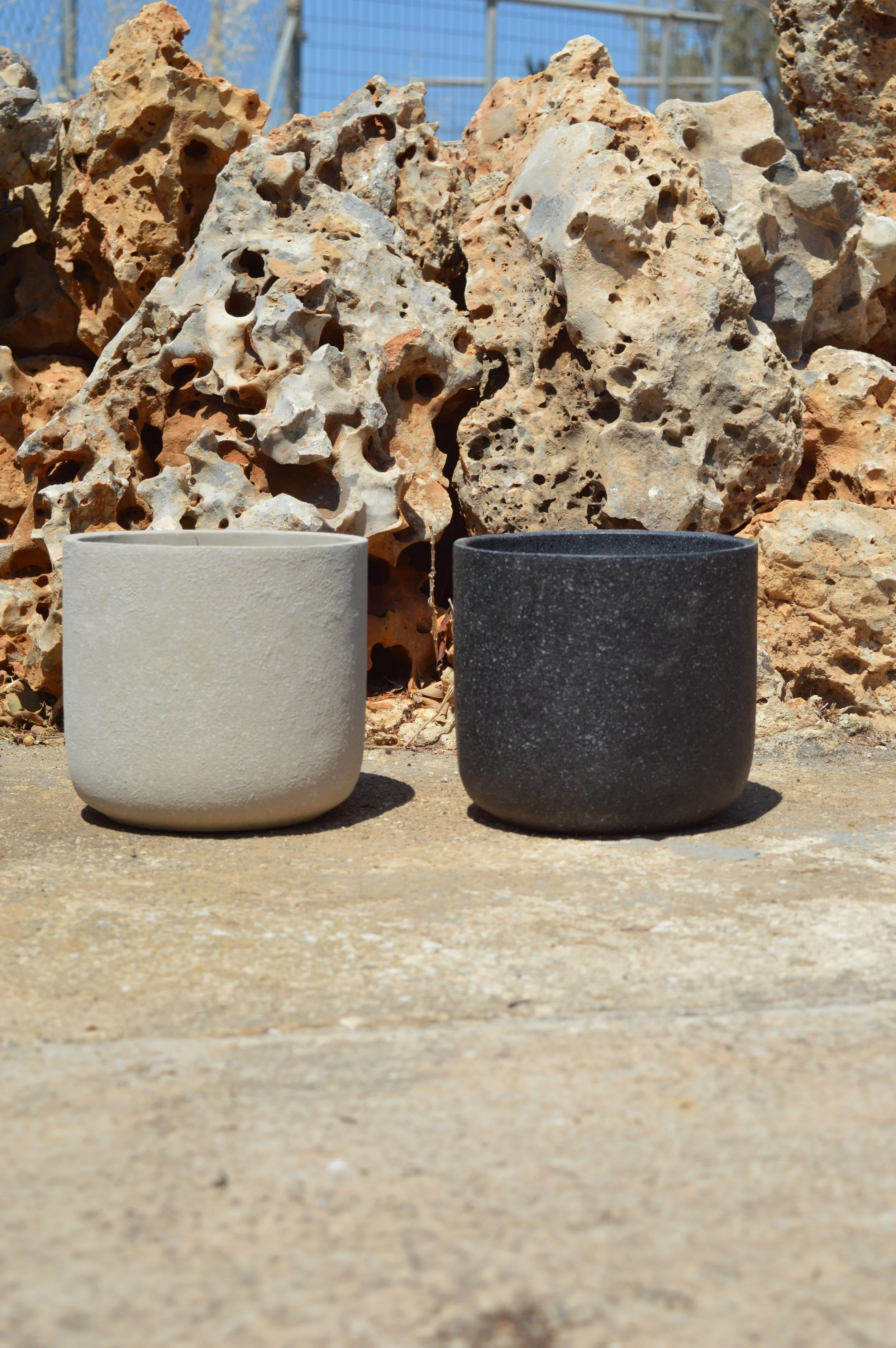 Close-up of two cylindrical planters, one white and one black, on a concrete surface with a rocky, sandy background.