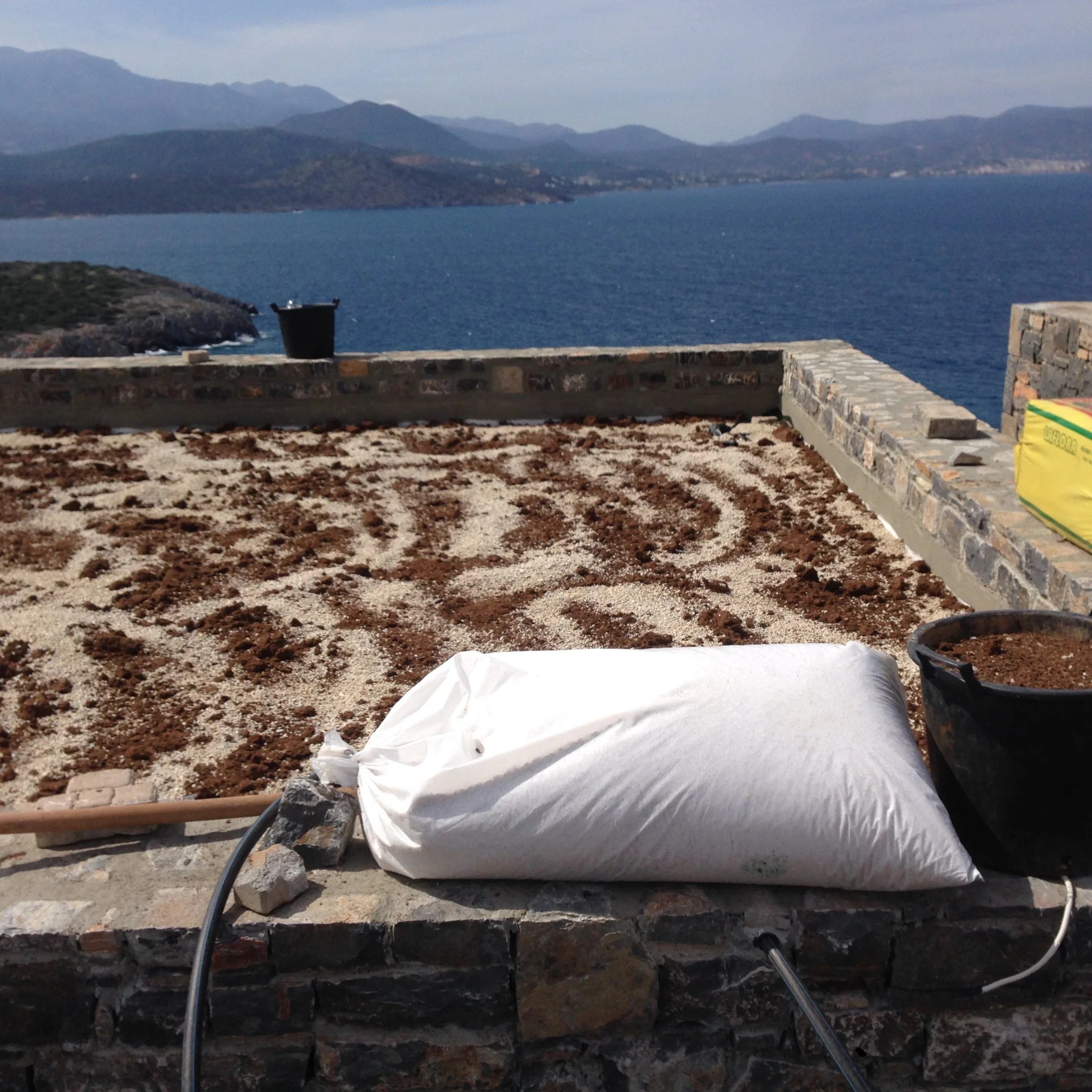 Photo of a scenic view overlooking a body of water with mountains in the background, taken from a stone patio or terrace with construction materials and tools in the foreground.