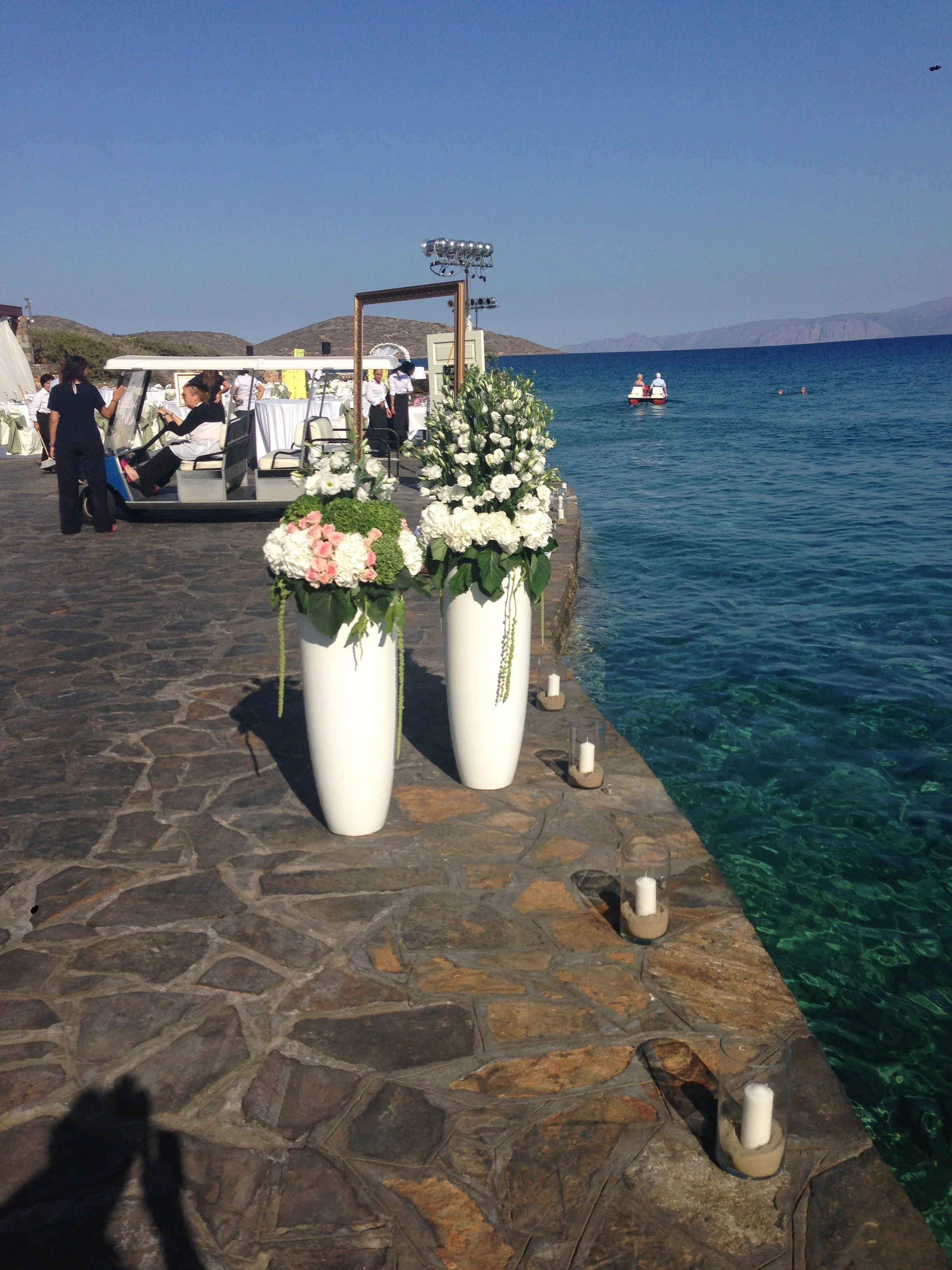 A waterfront scene with a stone patio, white flower arrangements in tall vases, and a boat on the water. There are people dressed in white and black, with some seated on a golf cart and others standing near it. The background features a clear blue sk