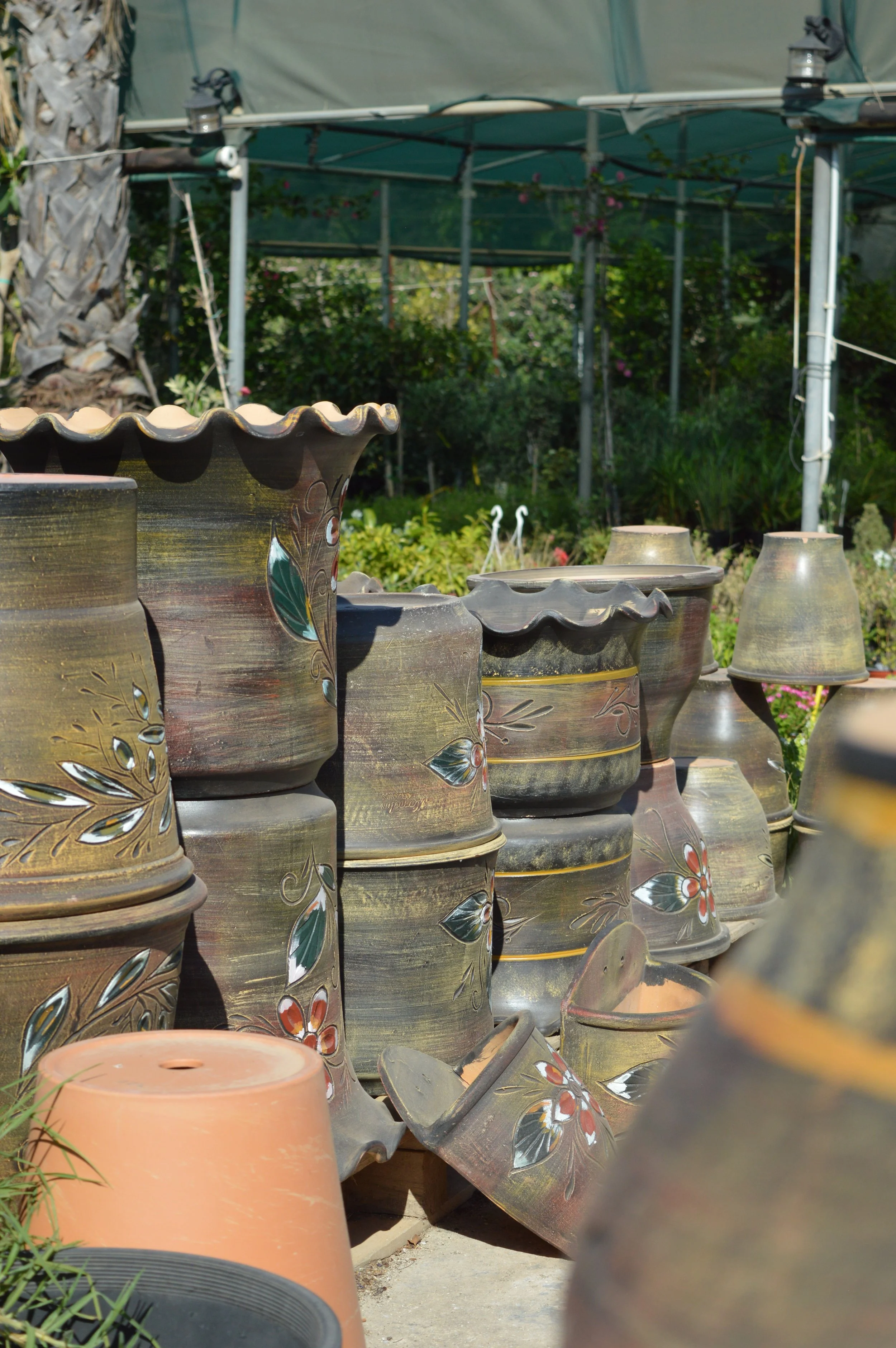 A collection of decorated ceramic flower pots and large planters outdoors, with a garden and greenhouse in the background.