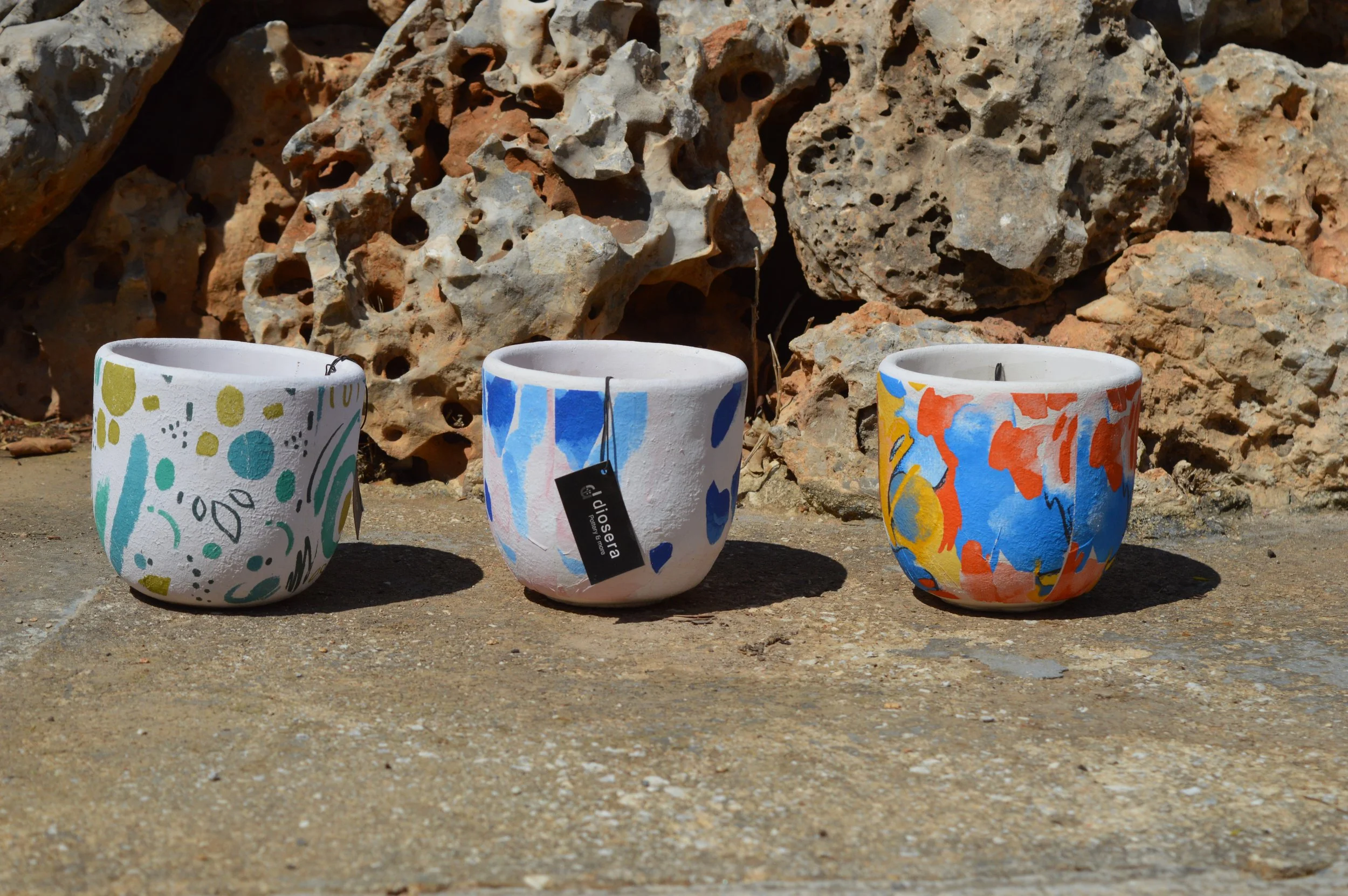 Three colorful, decorated ceramic flower pots placed on a concrete surface in front of a rocky background.