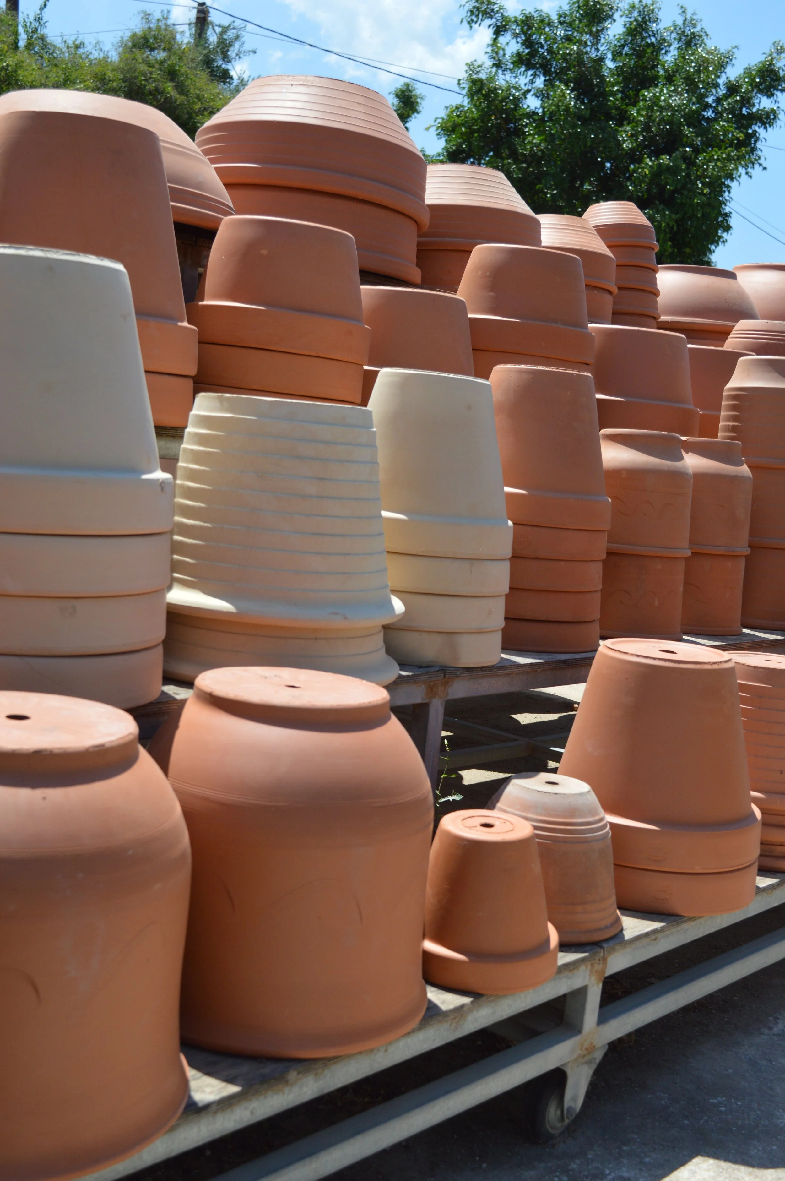 Multiple terracotta pots stacked and arranged outdoors on metal racks, with a background of trees and blue sky.