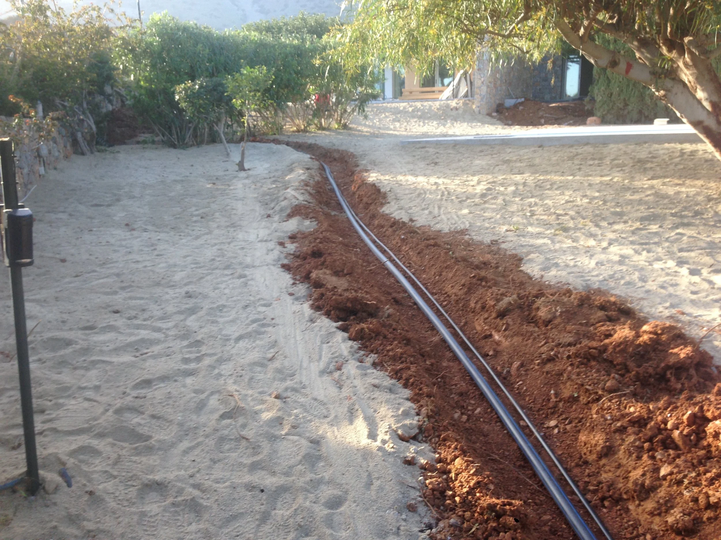 A construction site with a trench where underground piping is being installed, surrounded by dirt and gravel, with trees and a house in the background.