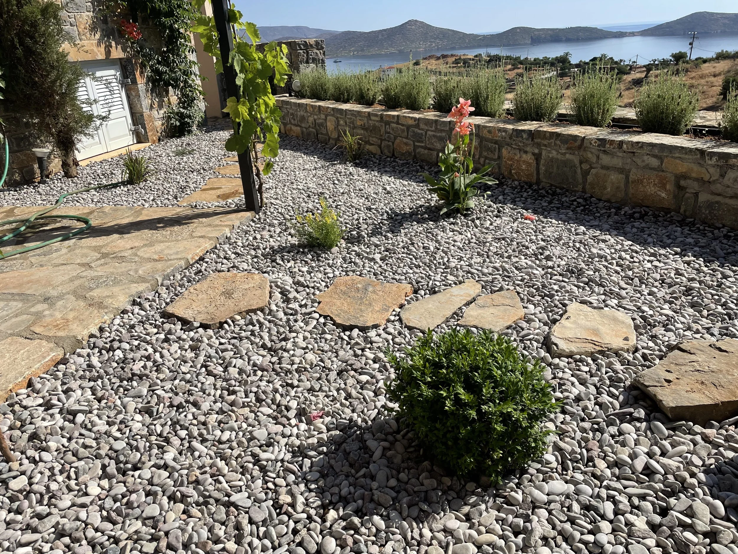 A stone patio with small stones, a garden hose, and a small plant in the foreground. There is a stone wall with lavender plants overlooking a body of water and distant hills in the background.