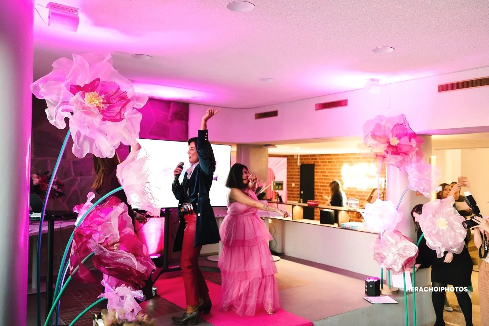 People dancing and singing at a pink-themed party, with large pink flower decorations, in a room with pink lighting and a bar area in the background.