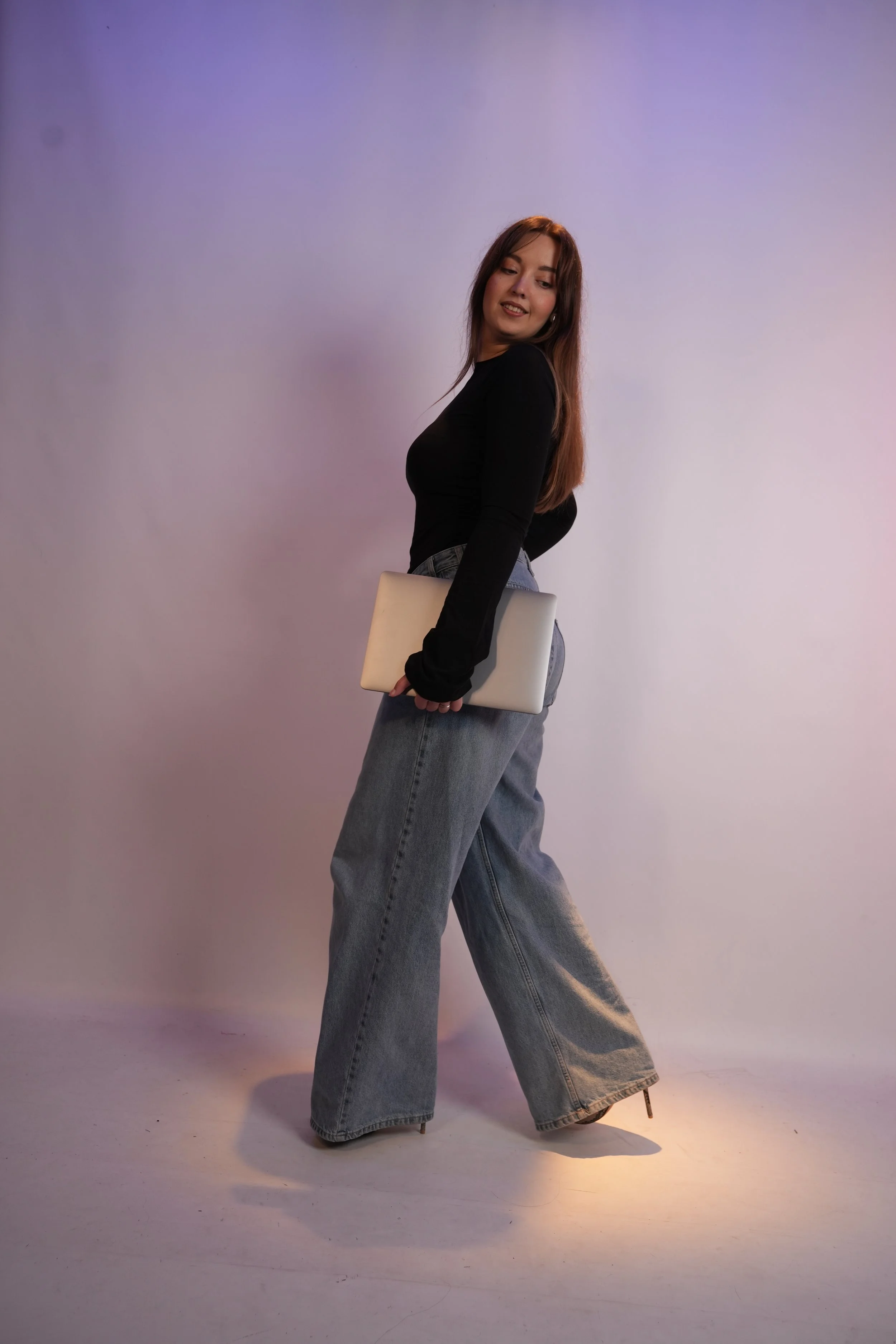 A young woman with long brown hair, wearing a black long-sleeve top and wide-leg jeans, stands sideways holding a closed laptop behind her back, with a purple lighting background.