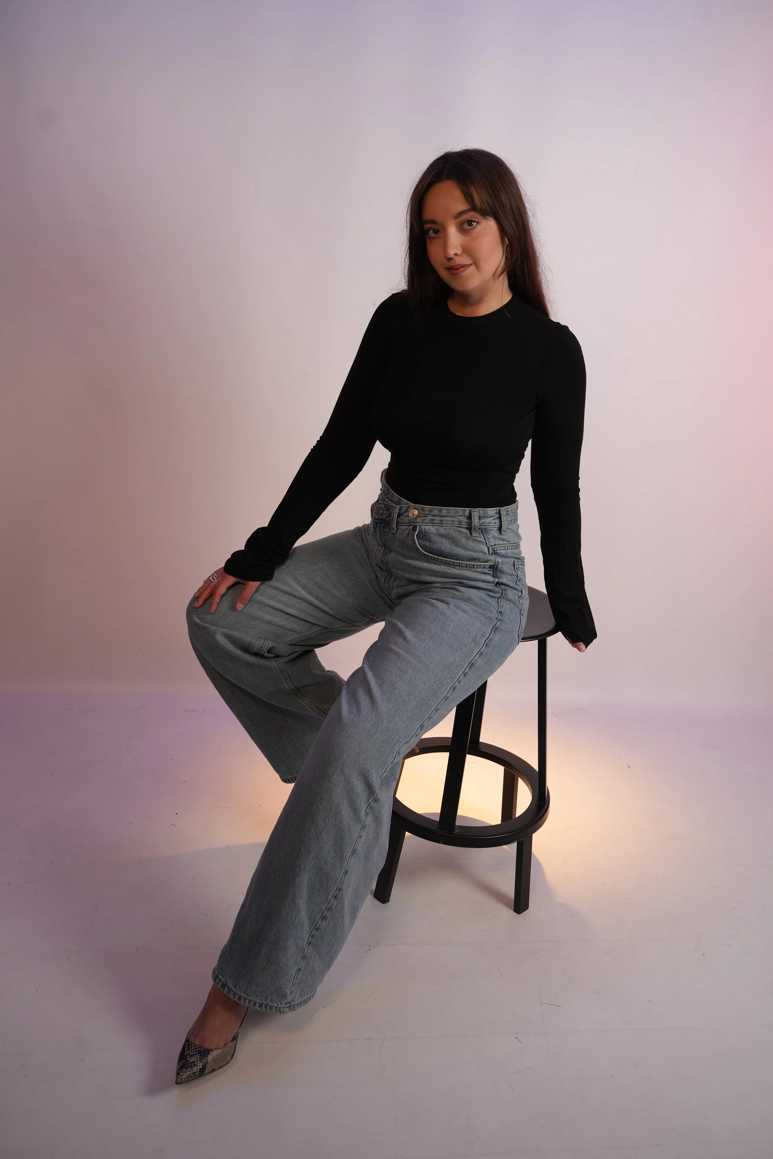 A young woman with long dark hair sitting on a high black stool against a plain white background. She is wearing a black long-sleeve shirt, light blue jeans, and pointed high heels with a snakeskin pattern. She has a confident expression and gazes directly at the camera.