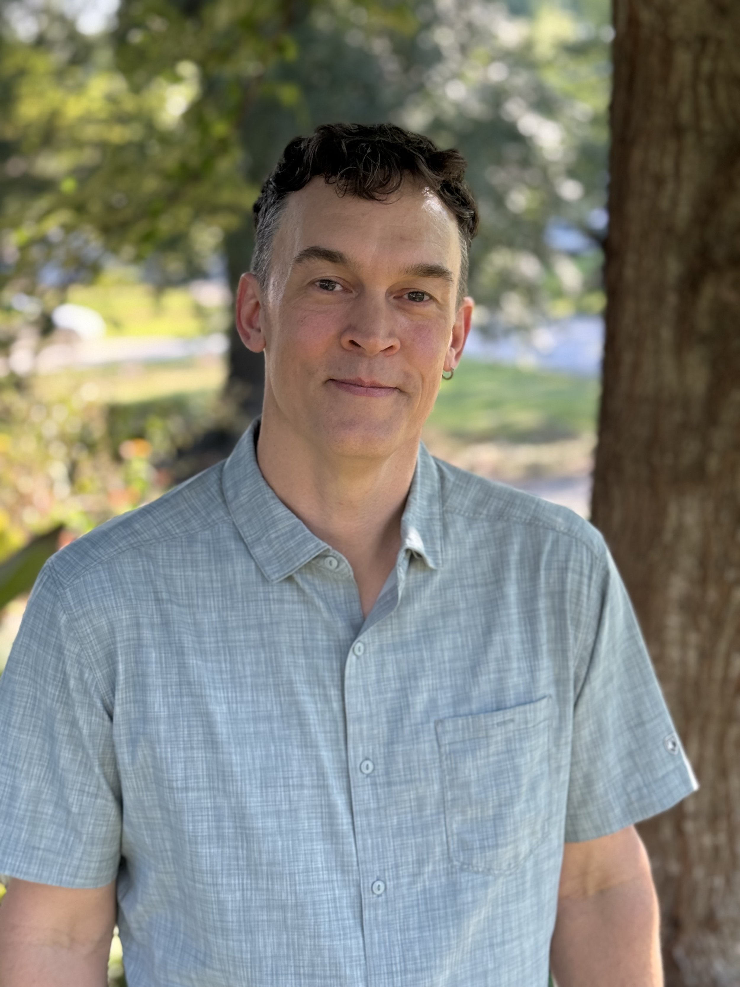 Close-up of a man wearing a light blue checkered shirt standing outdoors near a tree, with blurred greenery in the background.