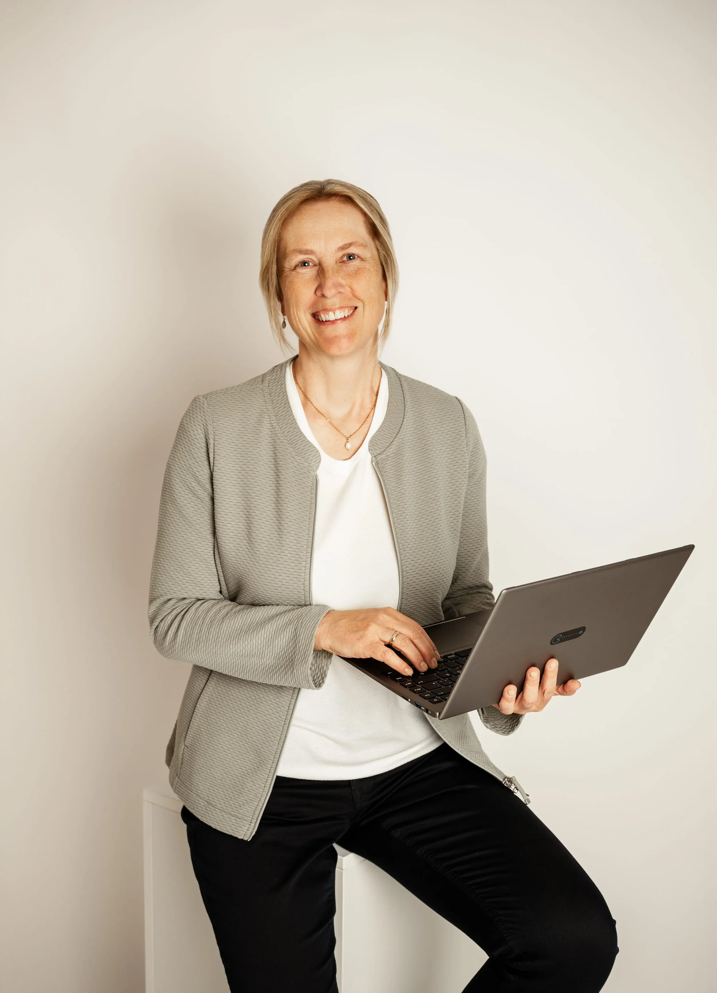 Eine lächelnde Frau mit kurzen blonden Haaren hält einen Laptop in der Hand und steht vor einer weißen Wand.
