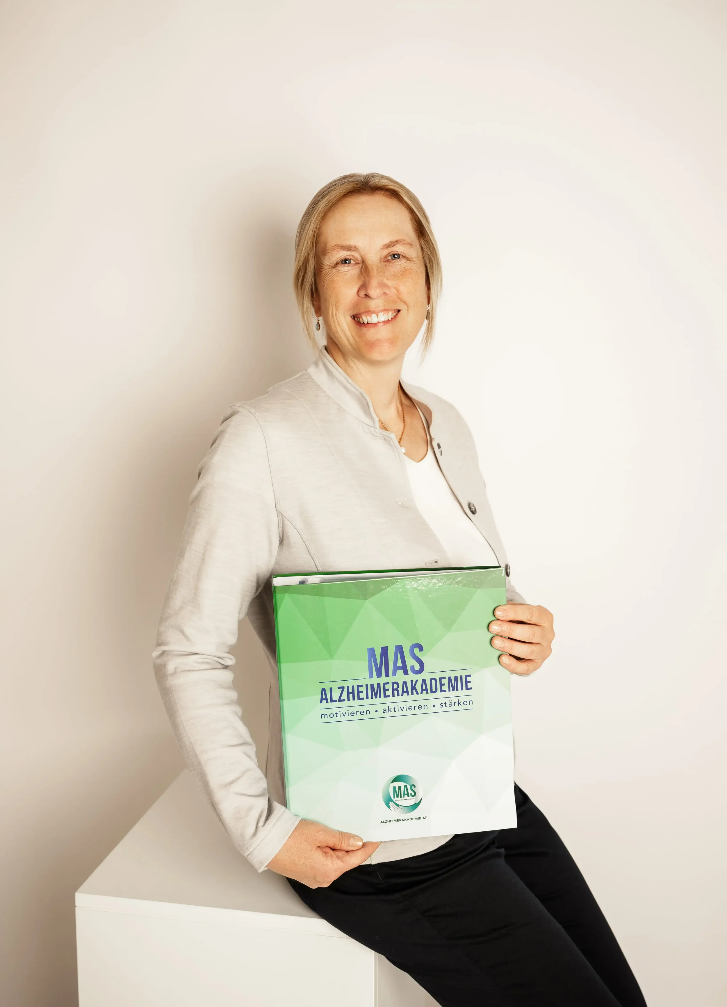 Eine lachende Frau hält einen Ordner mit dem Titel "MAS Alzheimerakademie" in den Händen, sie sitzt auf einer weißen Ablagefläche vor einem weißen Hintergrund.
