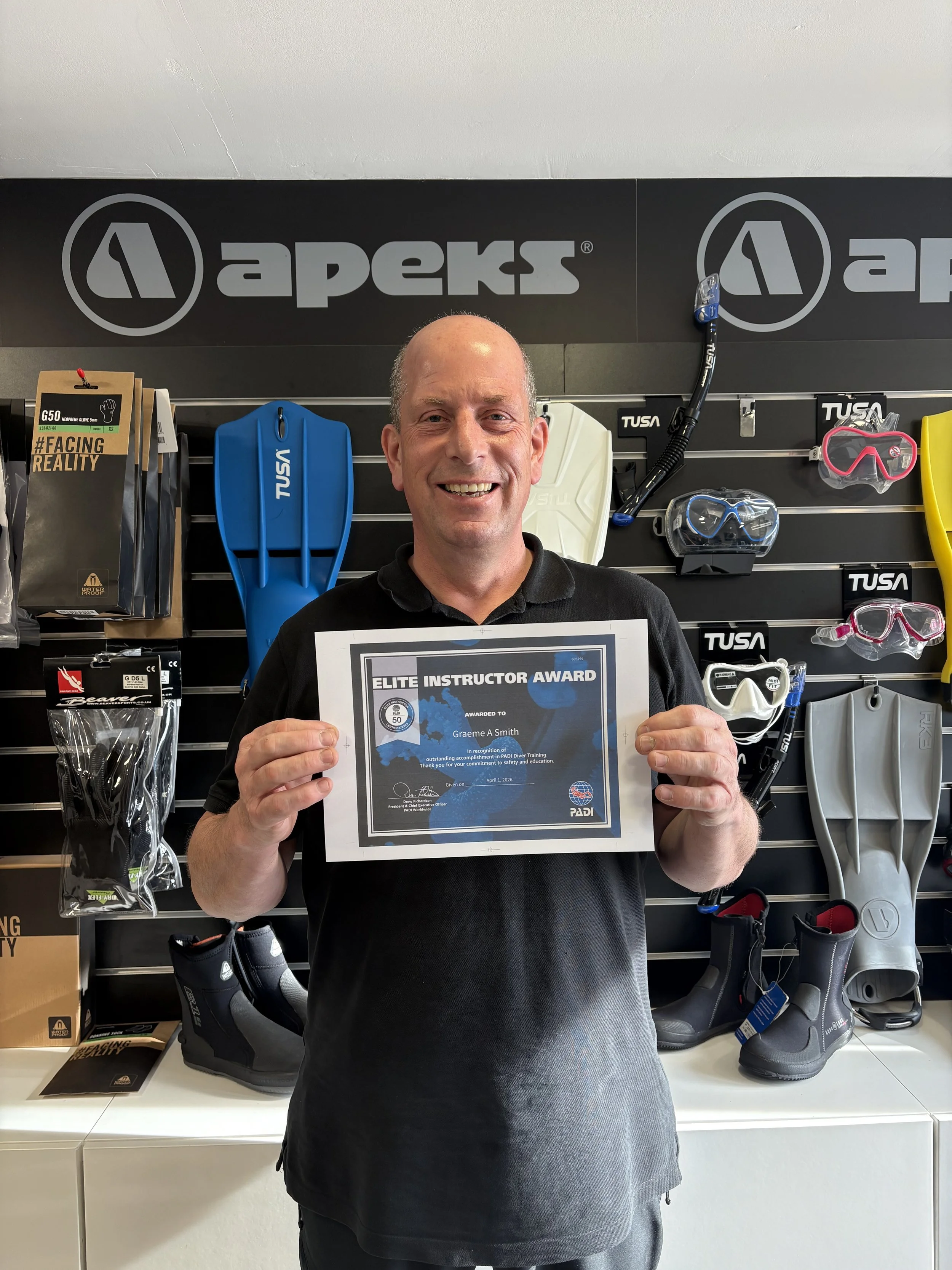 A man holding an award certificate in a retail store with outdoor and diving equipment on display.