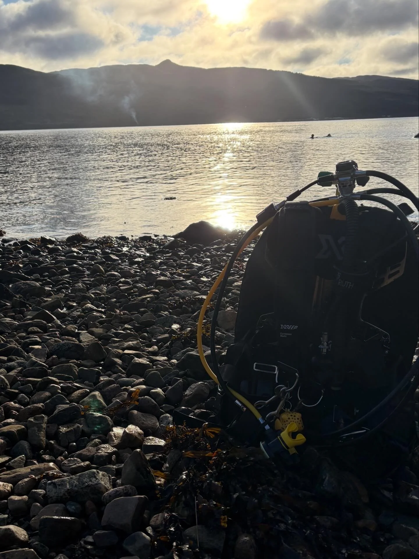 Scuba diving gear resting on rocky shoreline overlooking a lake with mountains in the background and the sun setting or rising.