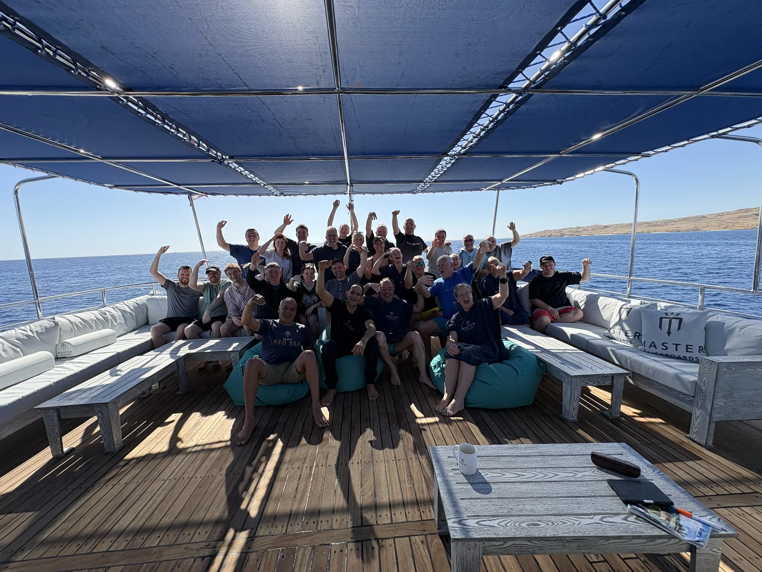 Group of people on a boat deck under a blue canopy, raising their arms and smiling, with ocean and distant land in the background.