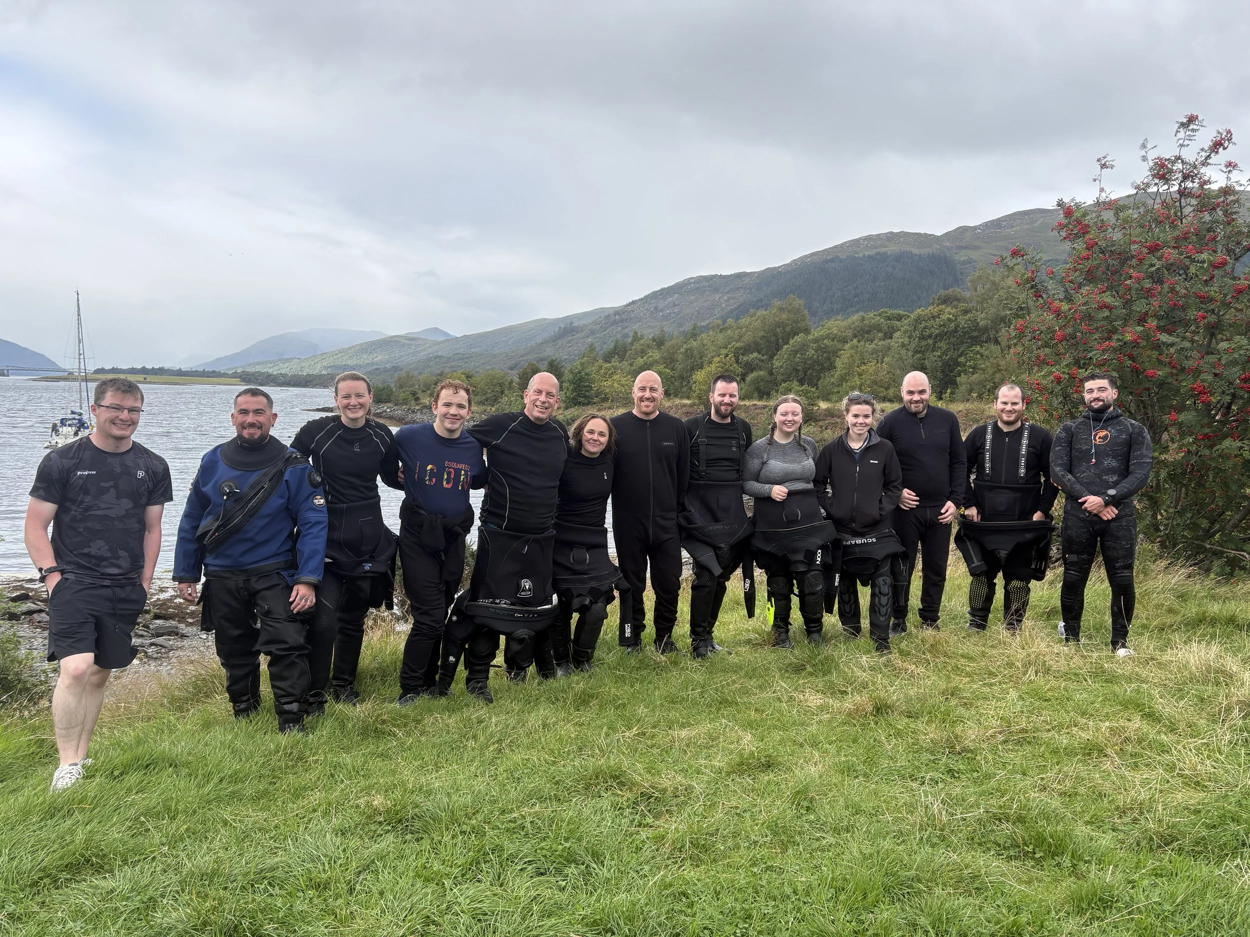 Group of ten people standing outdoors on grassy area near water, with mountains, trees, and overcast sky in the background.