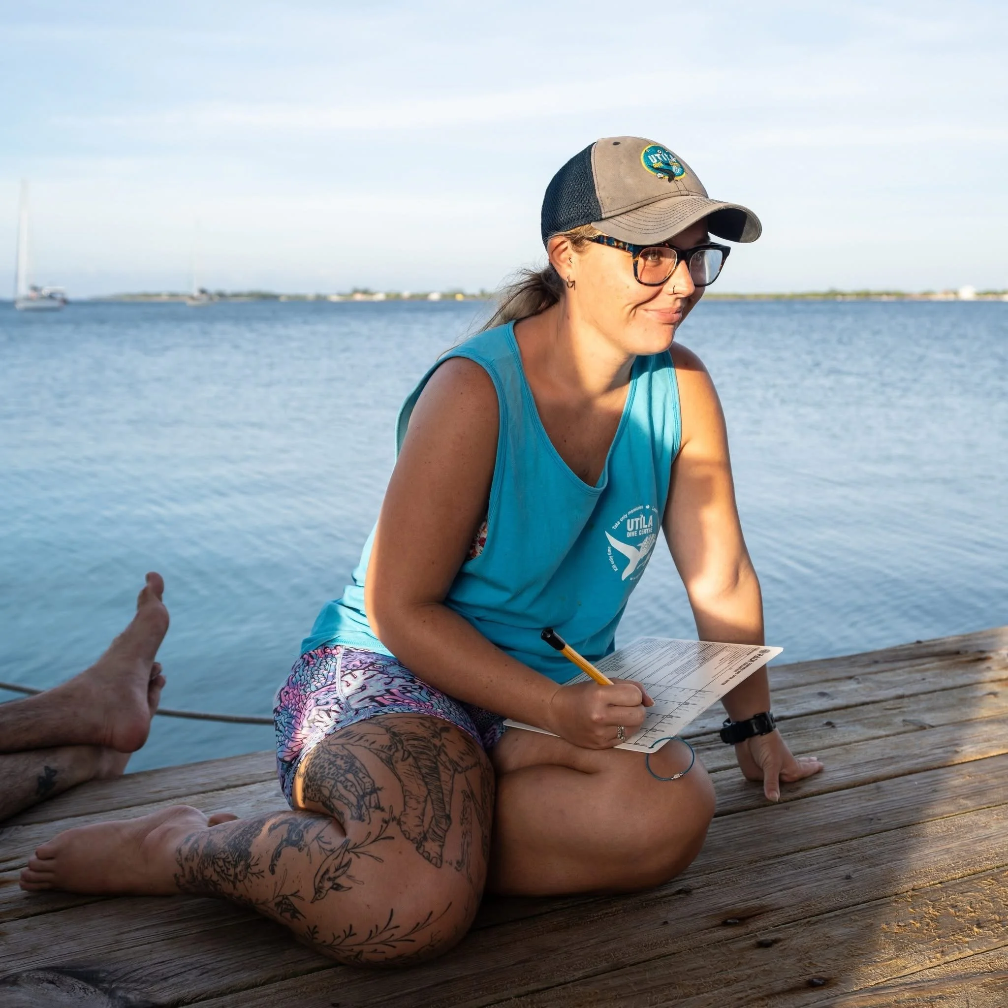 A woman with tattoos wearing a blue tank top, glasses, and a baseball cap, sitting on a wooden dock near water, writing on a notepad, with a hand and foot of another person visible in the foreground and a boat in the distance.