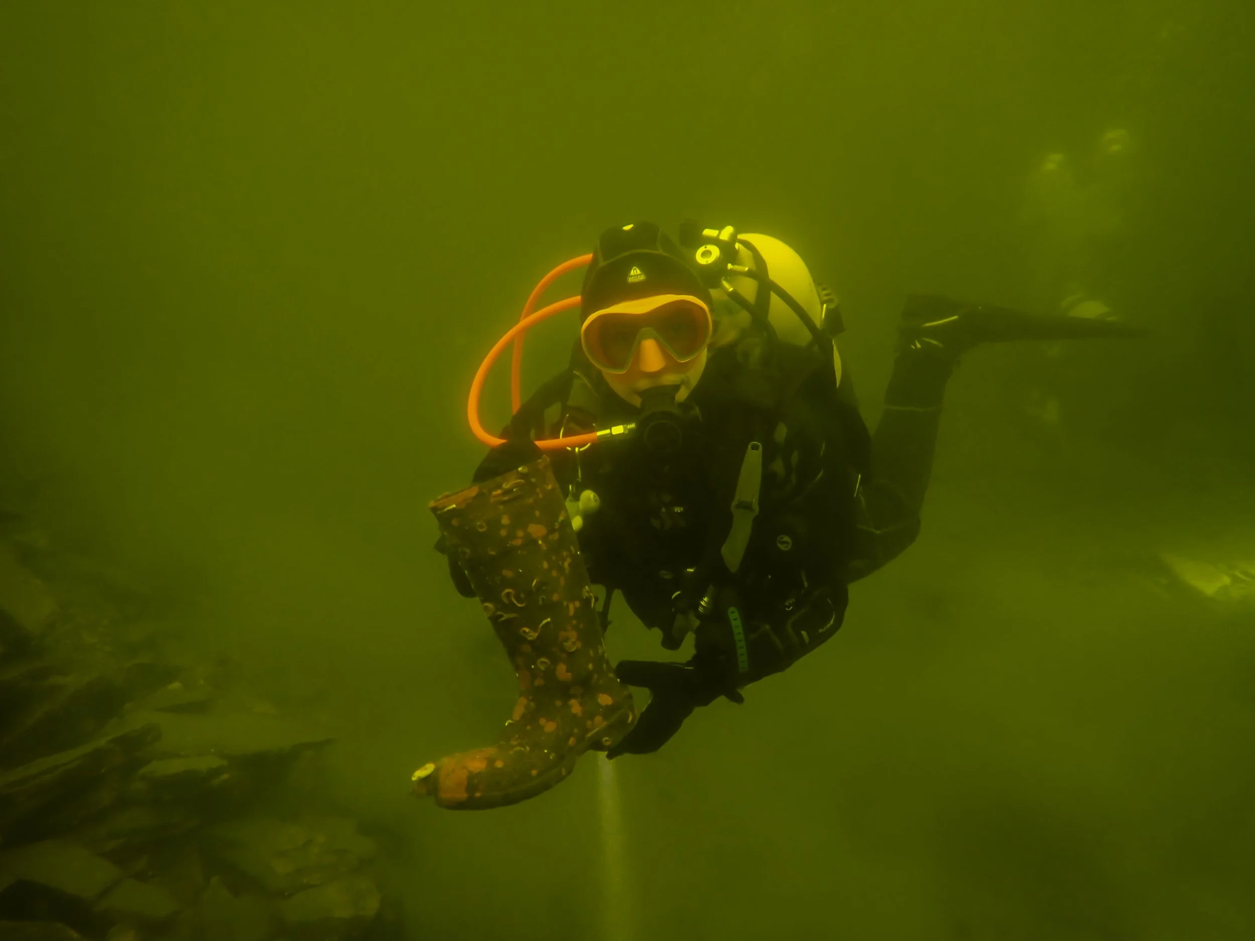 A scuba diver underwater, wearing a black wetsuit, yellow mask, and oxygen tank, swimming in greenish water.