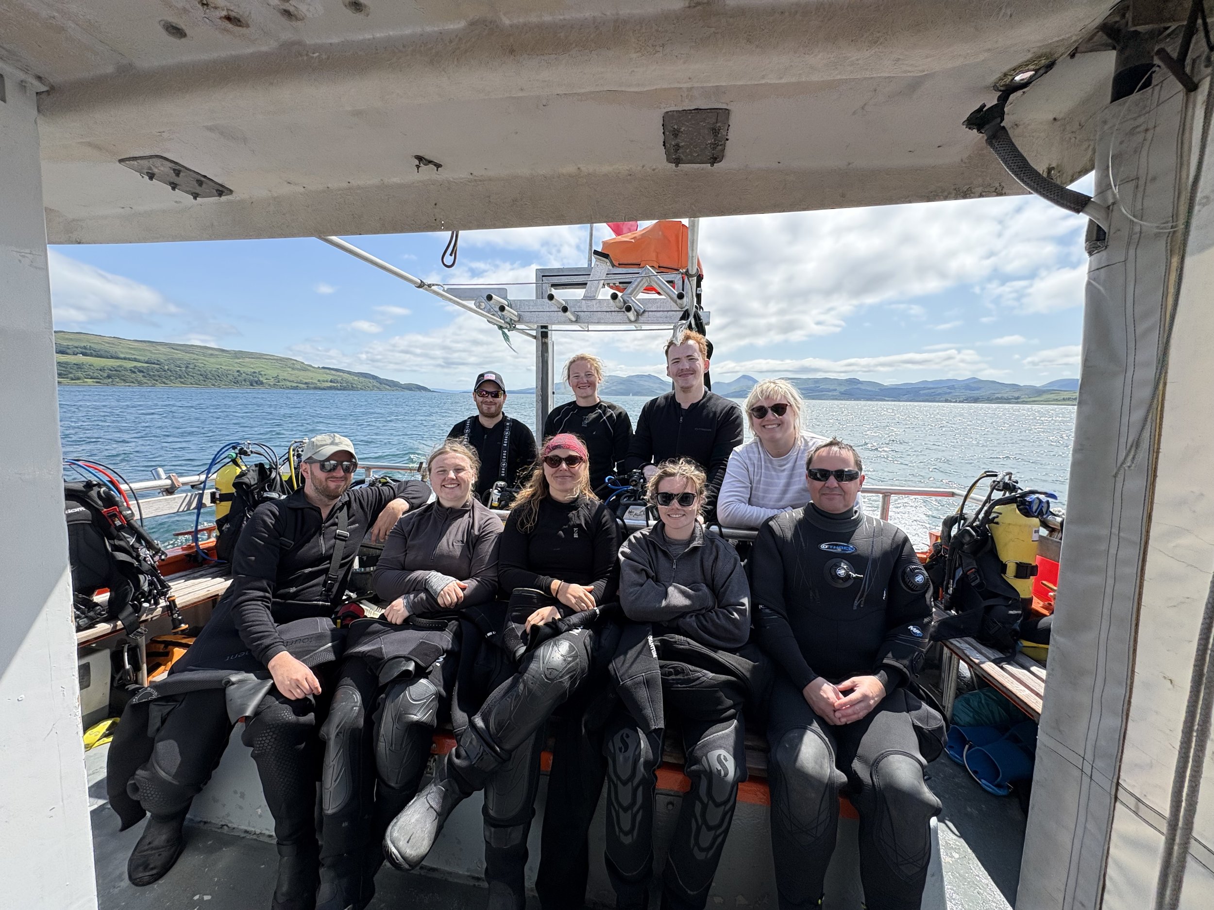 A group of nine people wearing scuba diving gear on a boat with a scenic water and hill view, possibly after a diving trip.