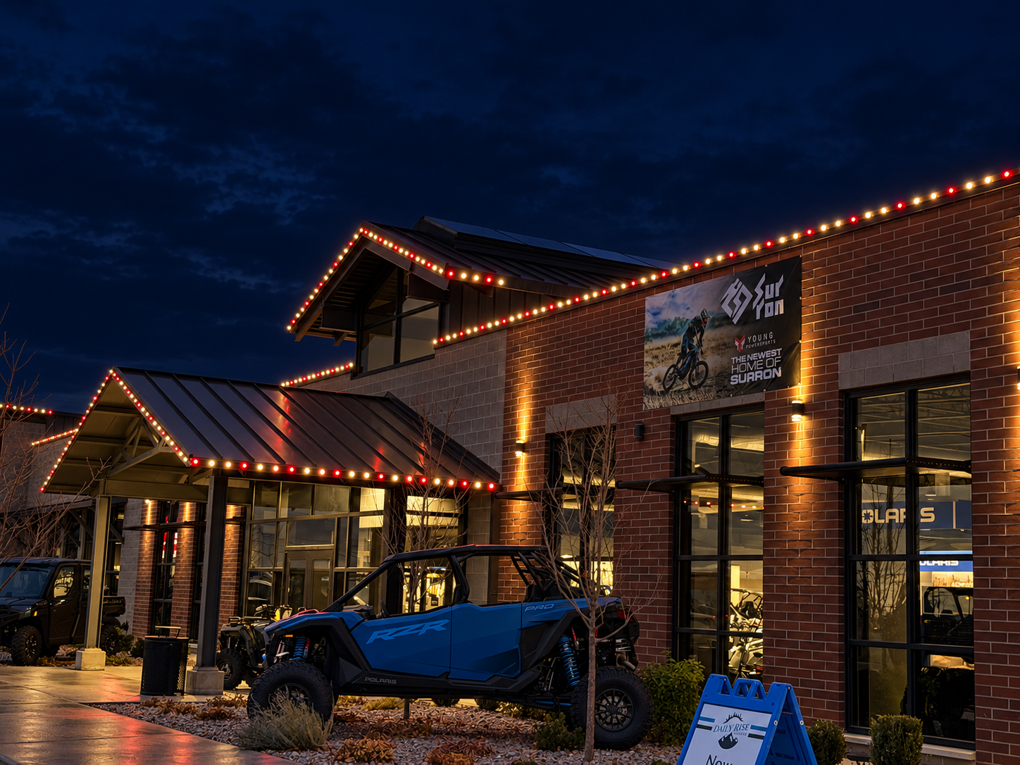 Exterior of a store at night decorated with red and white string lights, featuring a blue Polaris RZR side-by-side vehicle parked outside, with a brick facade and large glass windows, under a dark sky.