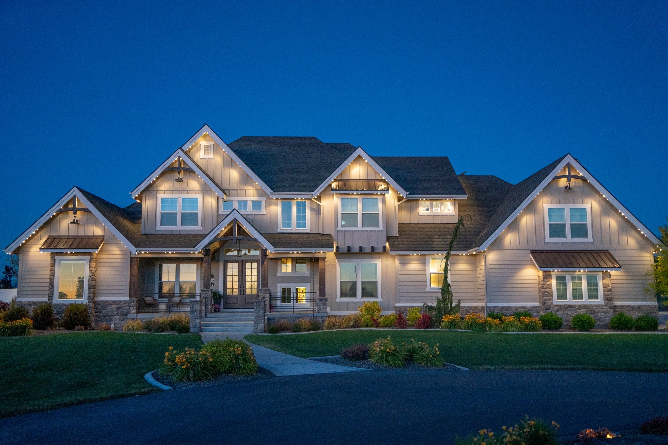 A modern house with multiple peaked roofs, illuminated with exterior lights, set against a twilight sky. There is a landscaped front yard with bushes and a curved pathway.