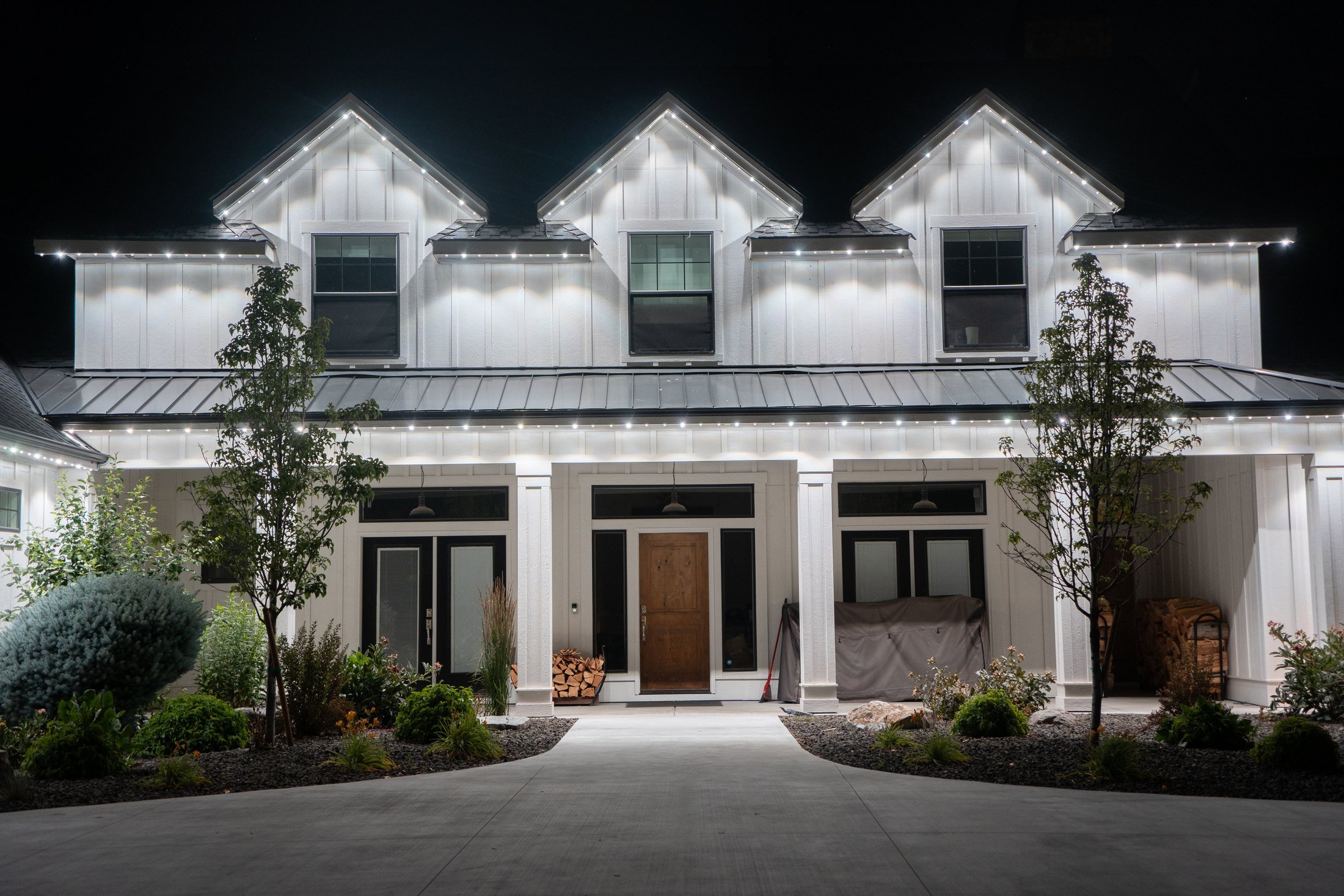A white house at night with exterior lights highlighting the architectural features, surrounded by a landscaped garden with trees and shrubs.