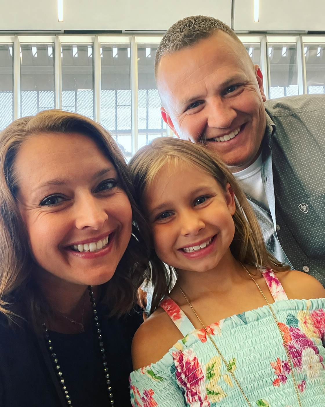 A smiling woman, a smiling young girl, and a smiling man are posing for a selfie inside a building with large windows.