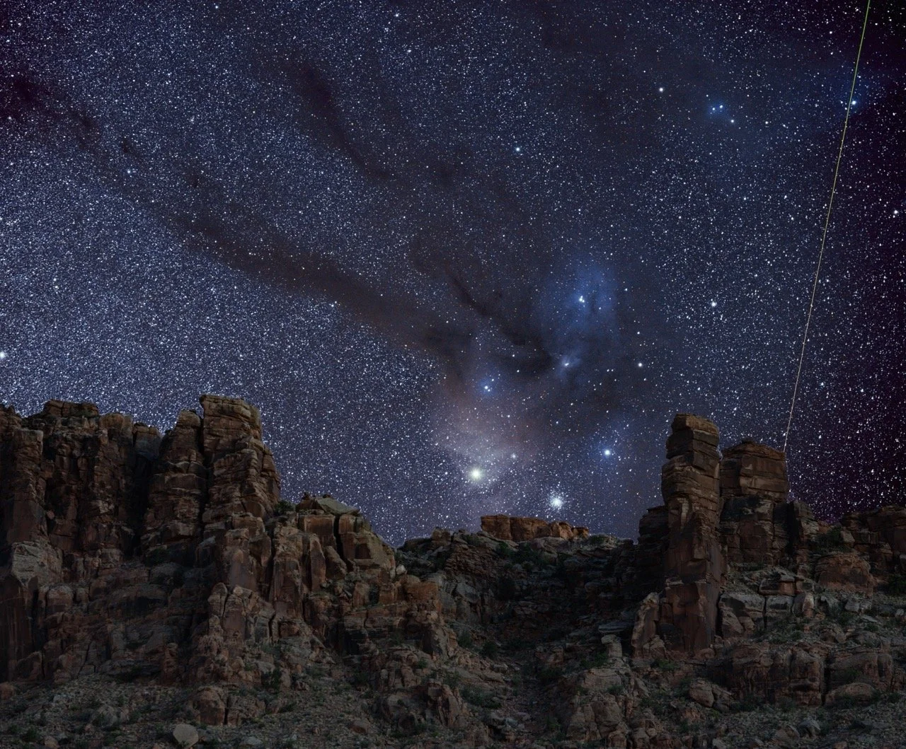 Night sky filled with stars above rocky desert landscape above hidden valley trailhead with large rock formations below the rho ophuchi complex and a piercing meteor from the perseids shooting through the atmosphere.