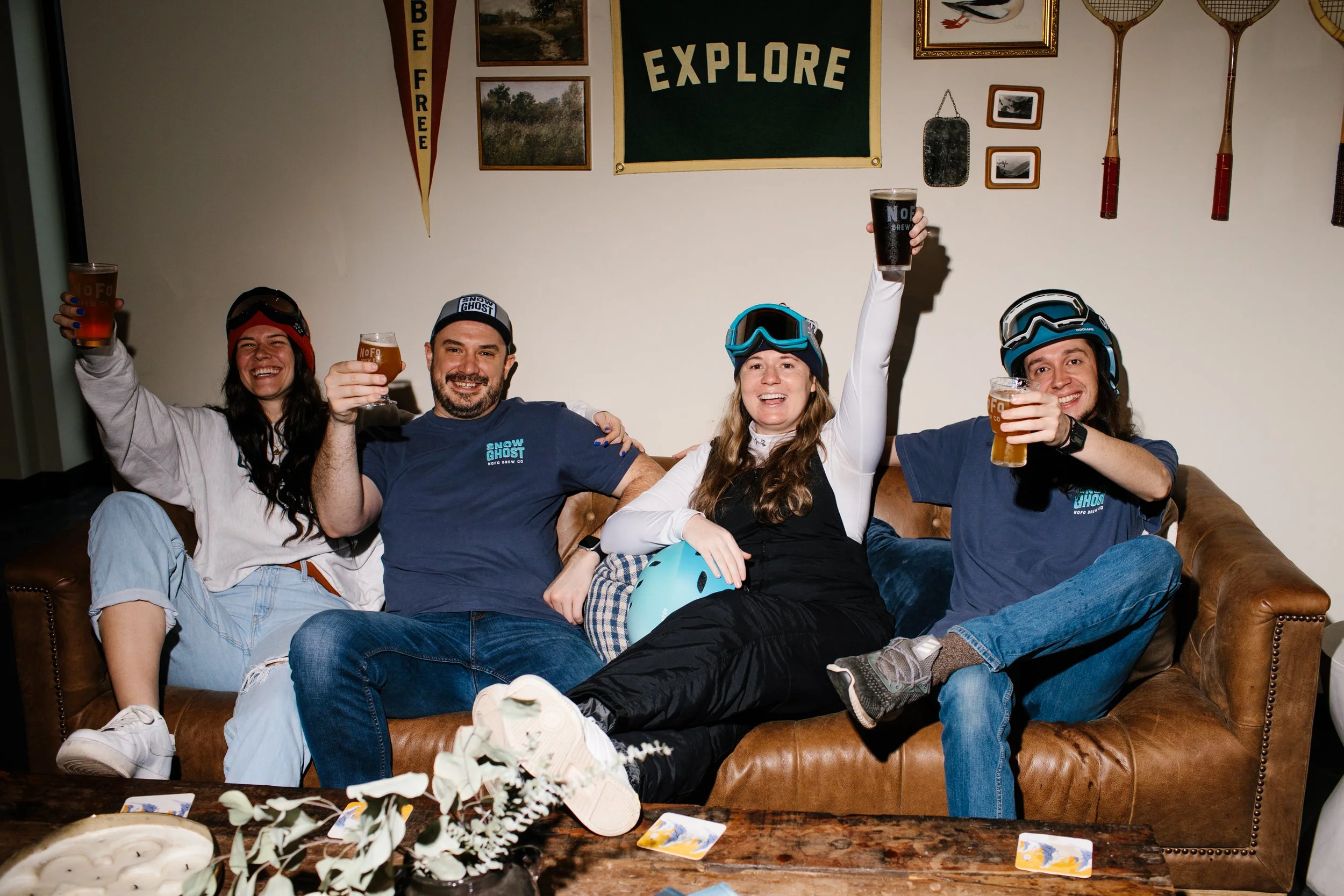 Four people sitting on a brown leather couch, raising glasses of beer and smiling. All are wearing helmets and outdoor gear, with mountain biking helmets on two of them. The background features a wall with pictures, a sign reading 'EXPLORE,' and fish