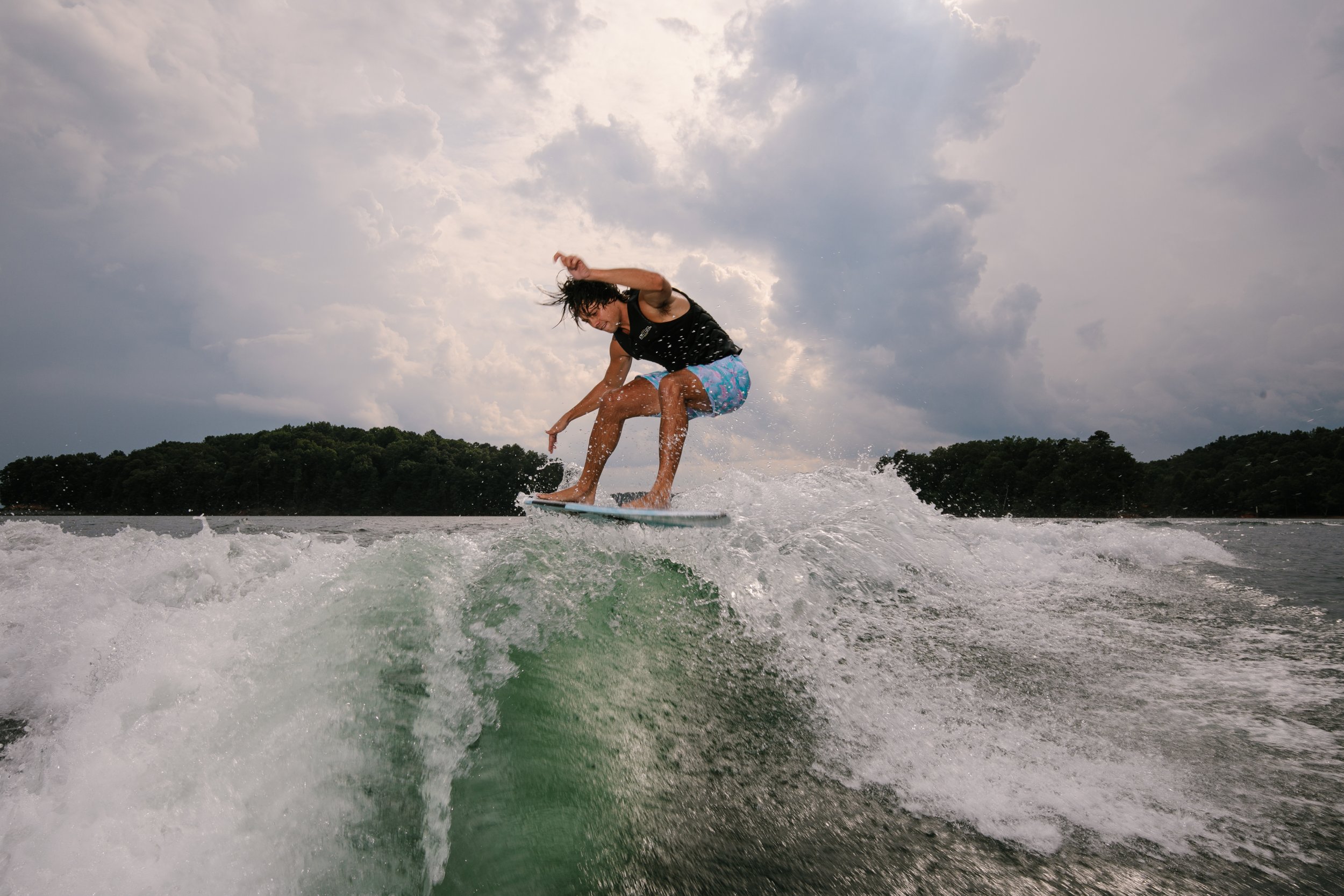 Person surfing on a wave on a body of water with trees and a cloudy sky in the background.
