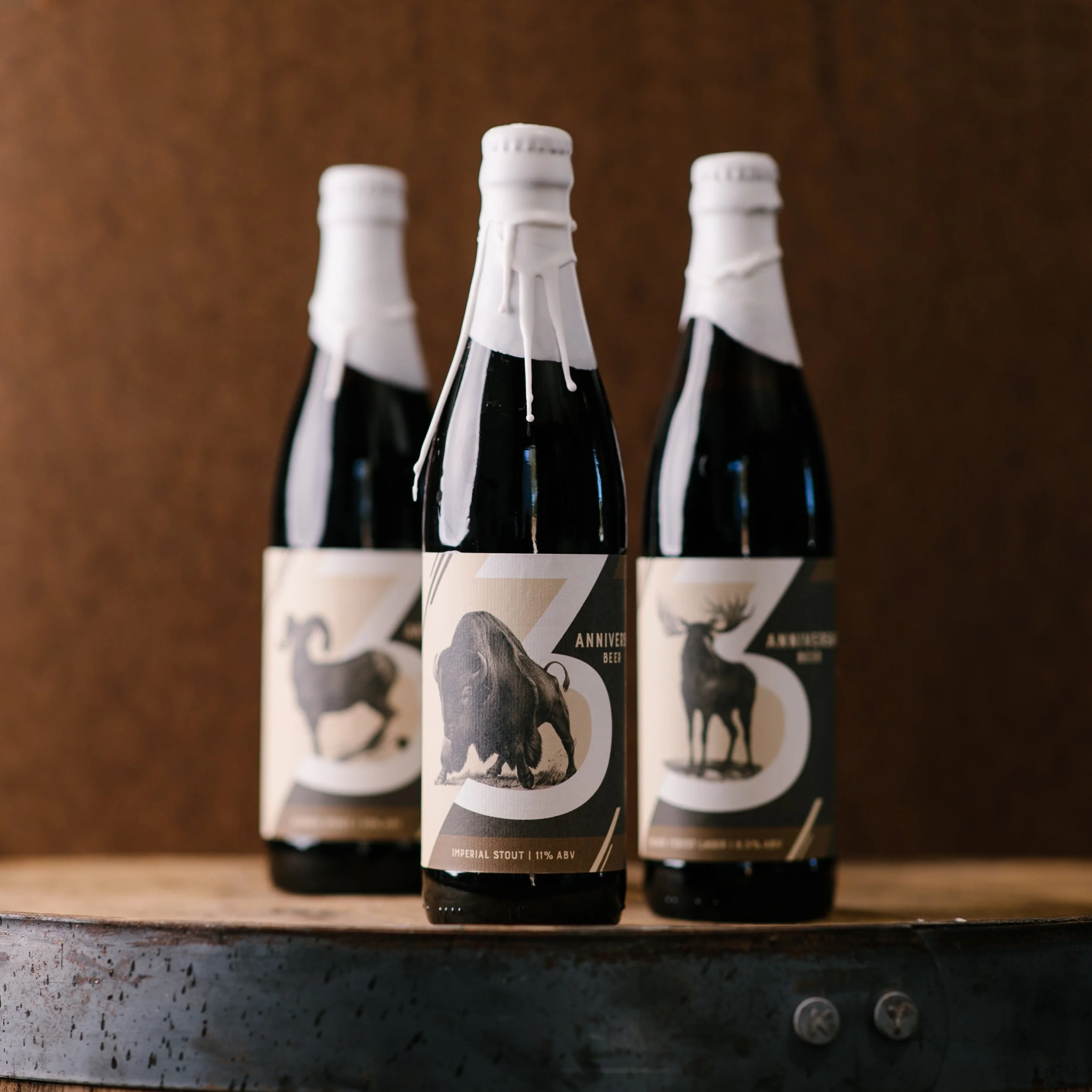 Close-up of a row of dark beer bottles with white bottle caps, placed on a wooden surface with a metallic background.