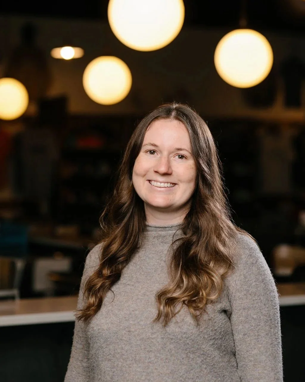 A woman with long wavy brown hair, smiling, wearing a light gray sweater, in a warmly lit indoor space with round hanging lights in the background.