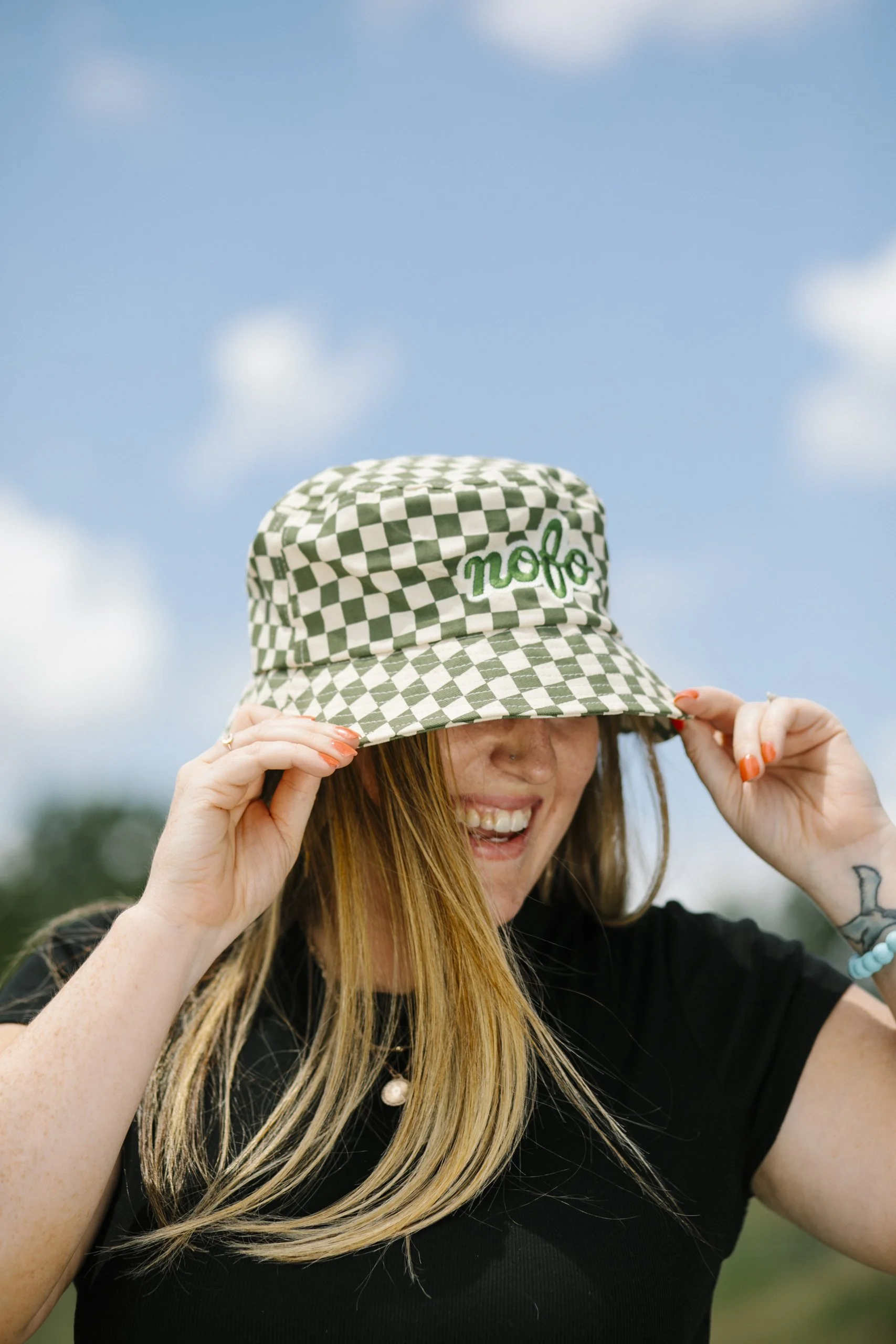 A woman smiling outdoors is adjusting a green and white checkered bucketembroidered on it in green. She has long blonde hair, a visible tattoo on her arm, and is wearing a black top with pearl and beaded accessories. The background shows a partly clo
