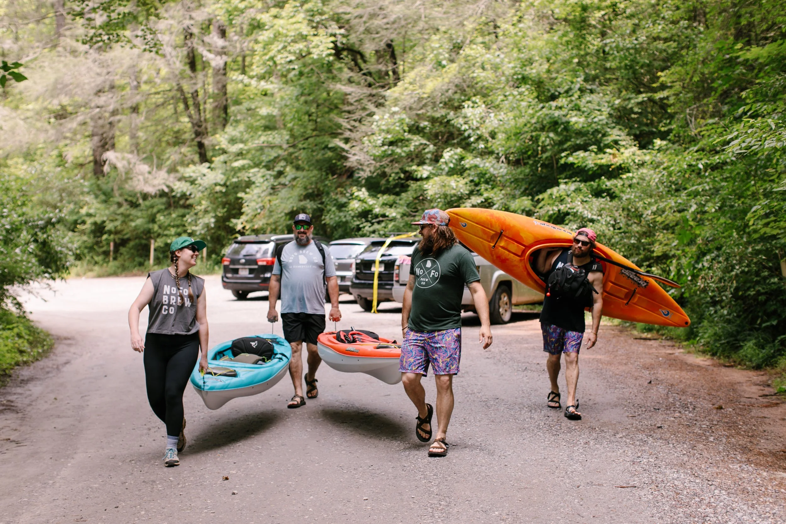 Group of four people carrying kayaks and gear, walking on a dirt path in a wooded area with parked cars in the background.