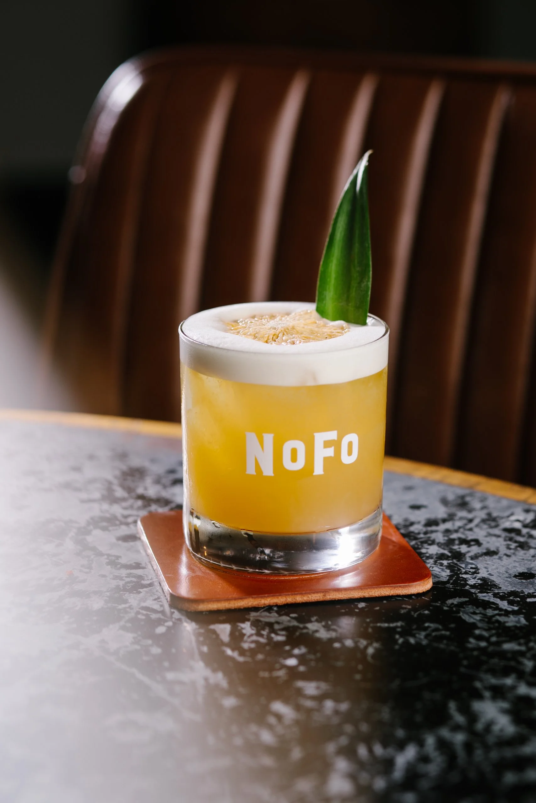 A yellow cocktail with foam on top, garnished with a green leaf, served in a glass with the words 'NoFo' on it, placed on a wooden coaster on a marble countertop.
