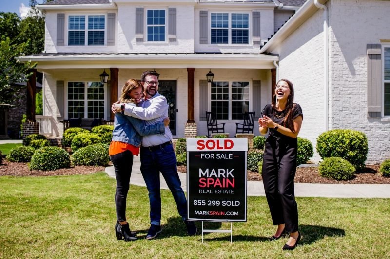 A couple embracing in front of a sold sign outside a house while a woman claps and smiles nearby.