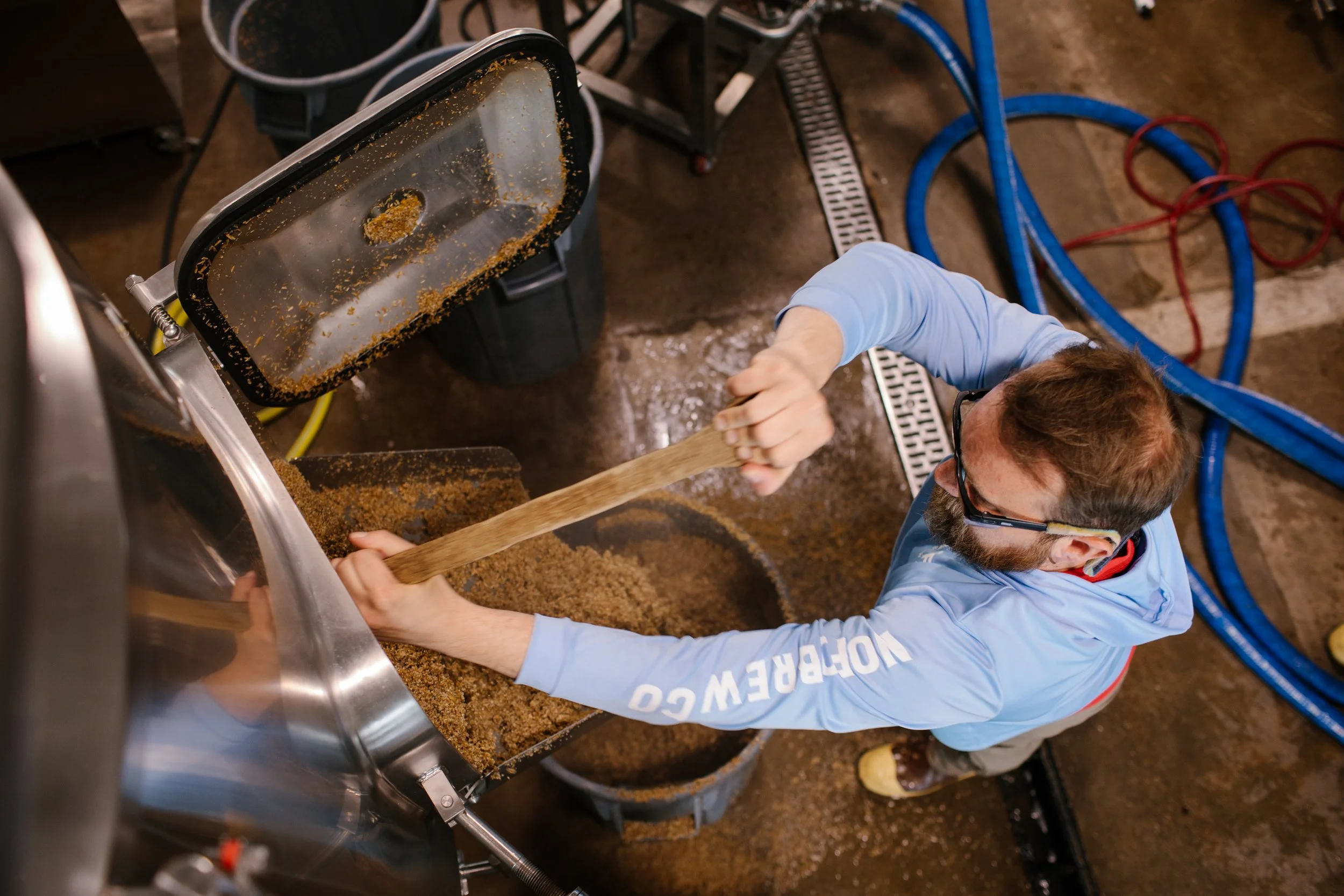 A person wearing safety glasses and a blue shirt is scooping a brown, granular substance from a large metal container with a wooden tool in an industrial or workshop setting.