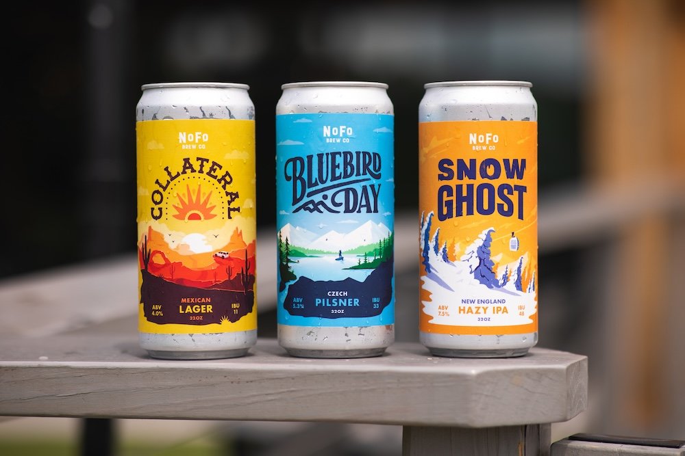 Three cans of craft beer displayed on a wooden surface, each with colorful labels featuring distinct outdoor scenic illustrations and text.