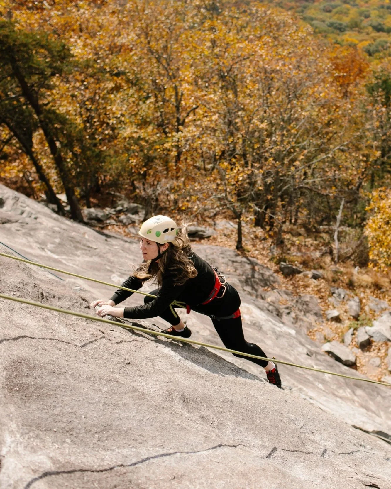 A woman wearing climbing gear and a helmet is climbing a steep rock face with a harness and rope, surrounded by autumn trees with yellow and orange leaves.