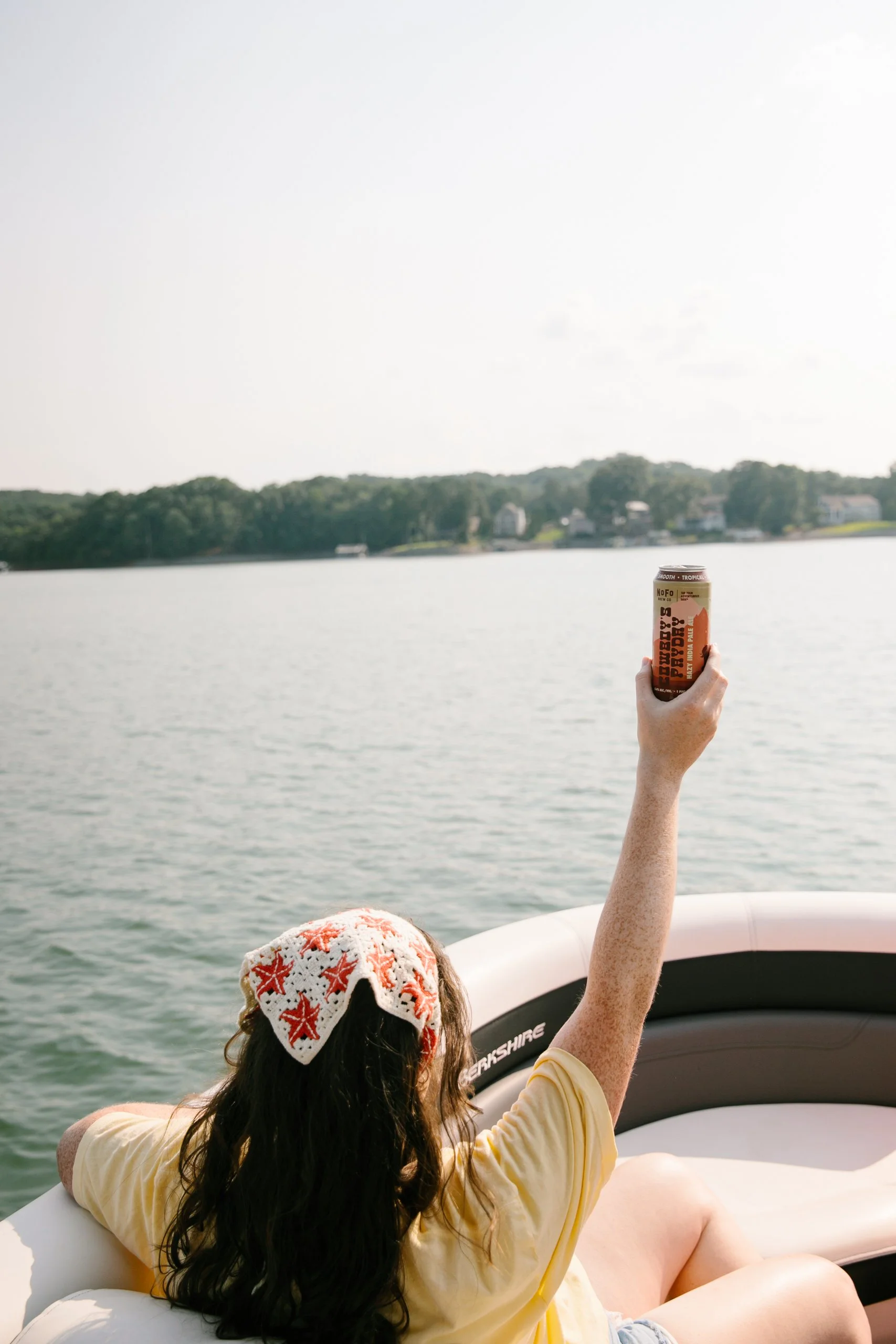 Person with long dark hair wearing a red and white star-patterned hat, holding up a can of beer while sitting on an inflatable boat on a lake with houses and trees in the background.