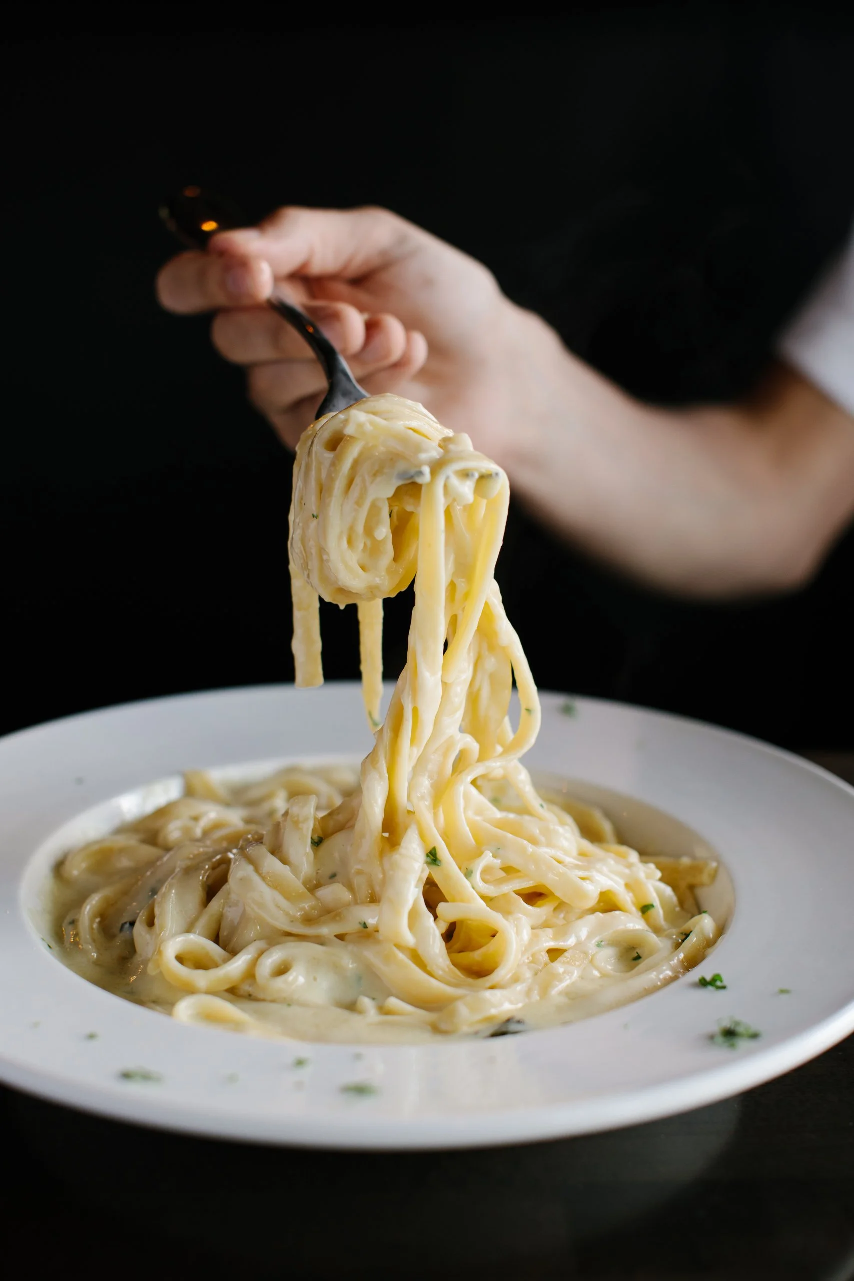 A plate of creamy Alfredo pasta with a fork lifting a serving, garnished with herbs, against a dark background.