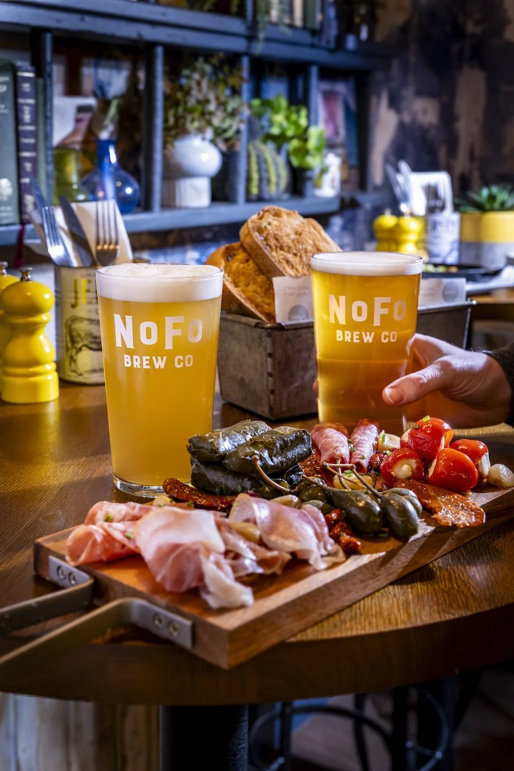 Two glasses of beer along with a charcuterie board with various meats, cheeses, and tomatoes, on a wooden table in a cozy, decorated bar or café setting.