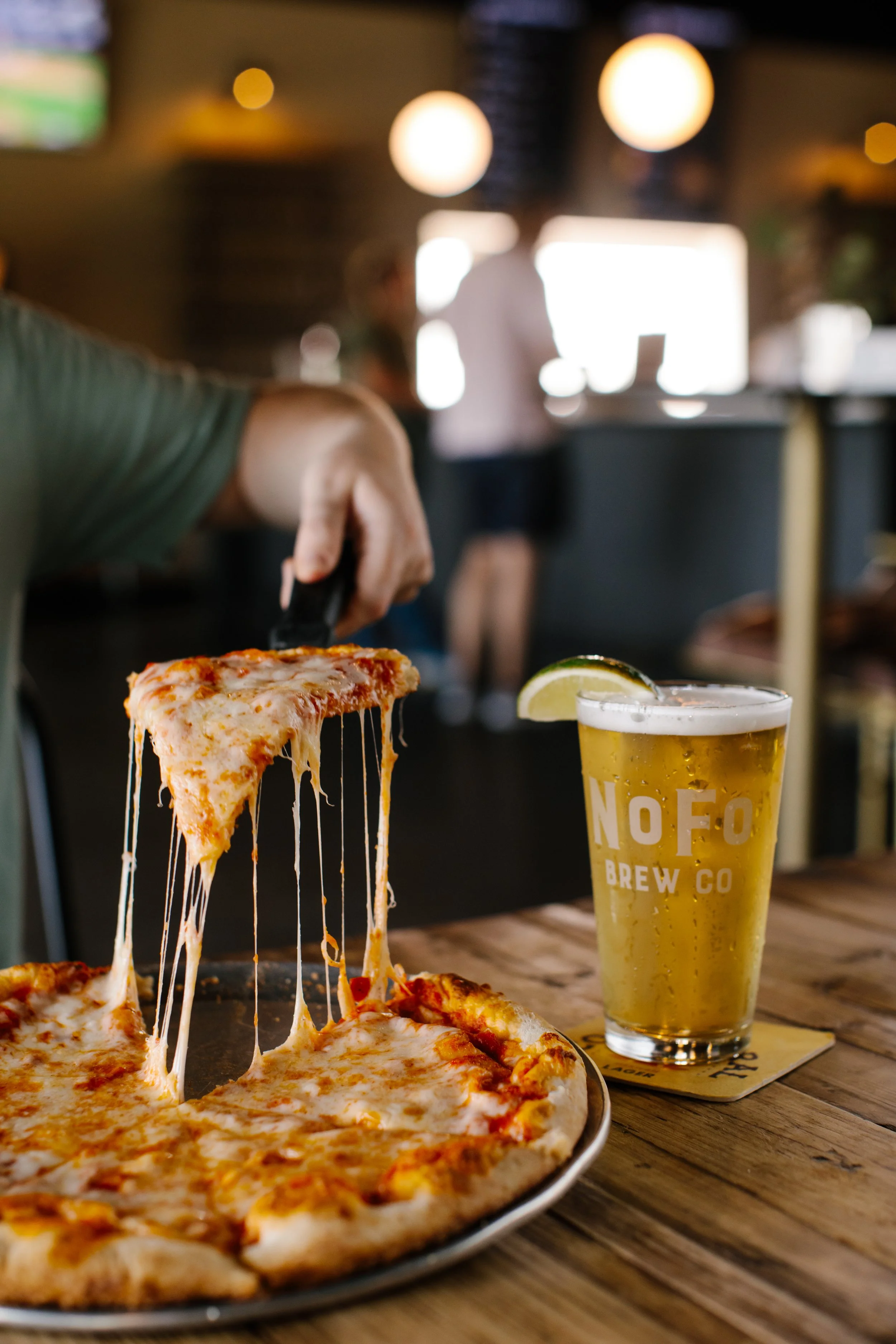 A slice of cheese pizza being lifted with melted cheese stretching, on a plate, beside a pint of beer garnished with a lemon wedge on a wooden table in a restaurant.
