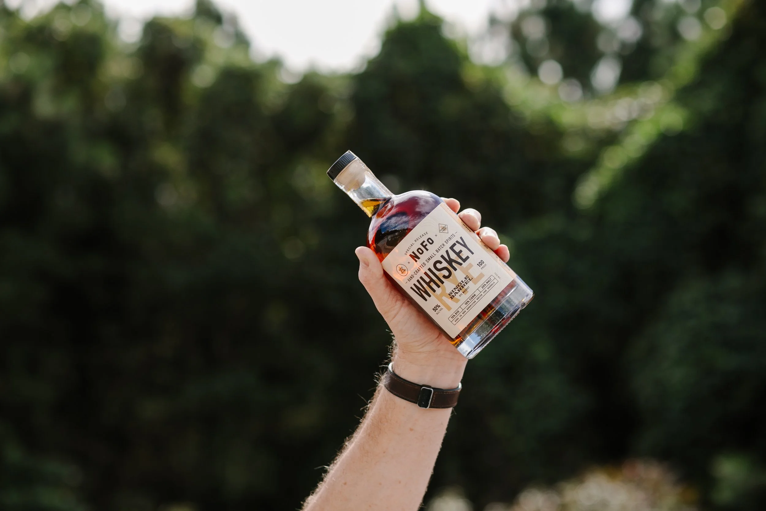 Person holding a bottle of whiskey outdoors with a blurred green forest background.