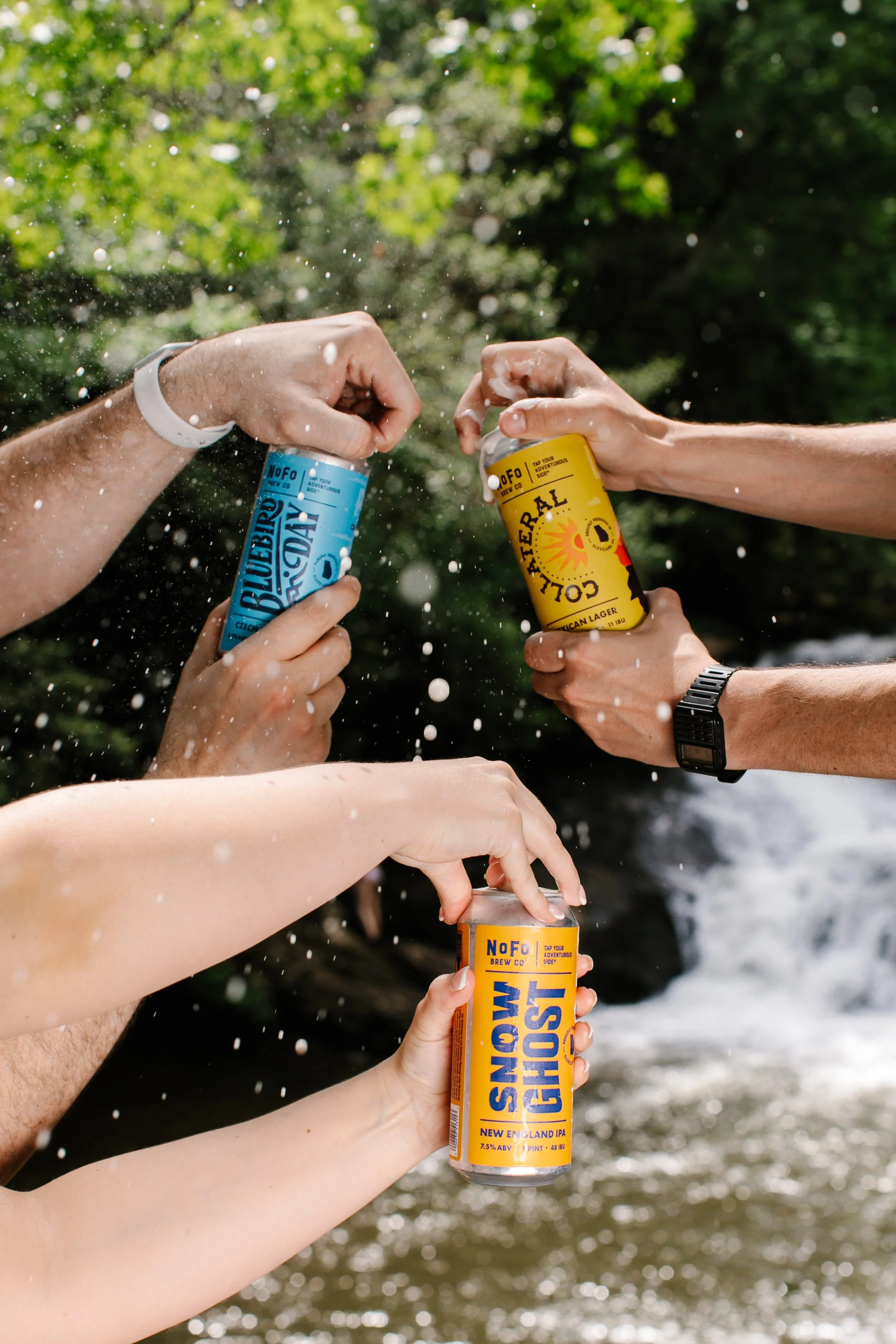 Multiple people holding cans of beer and popping the tops off in an outdoor setting with trees and water in the background.